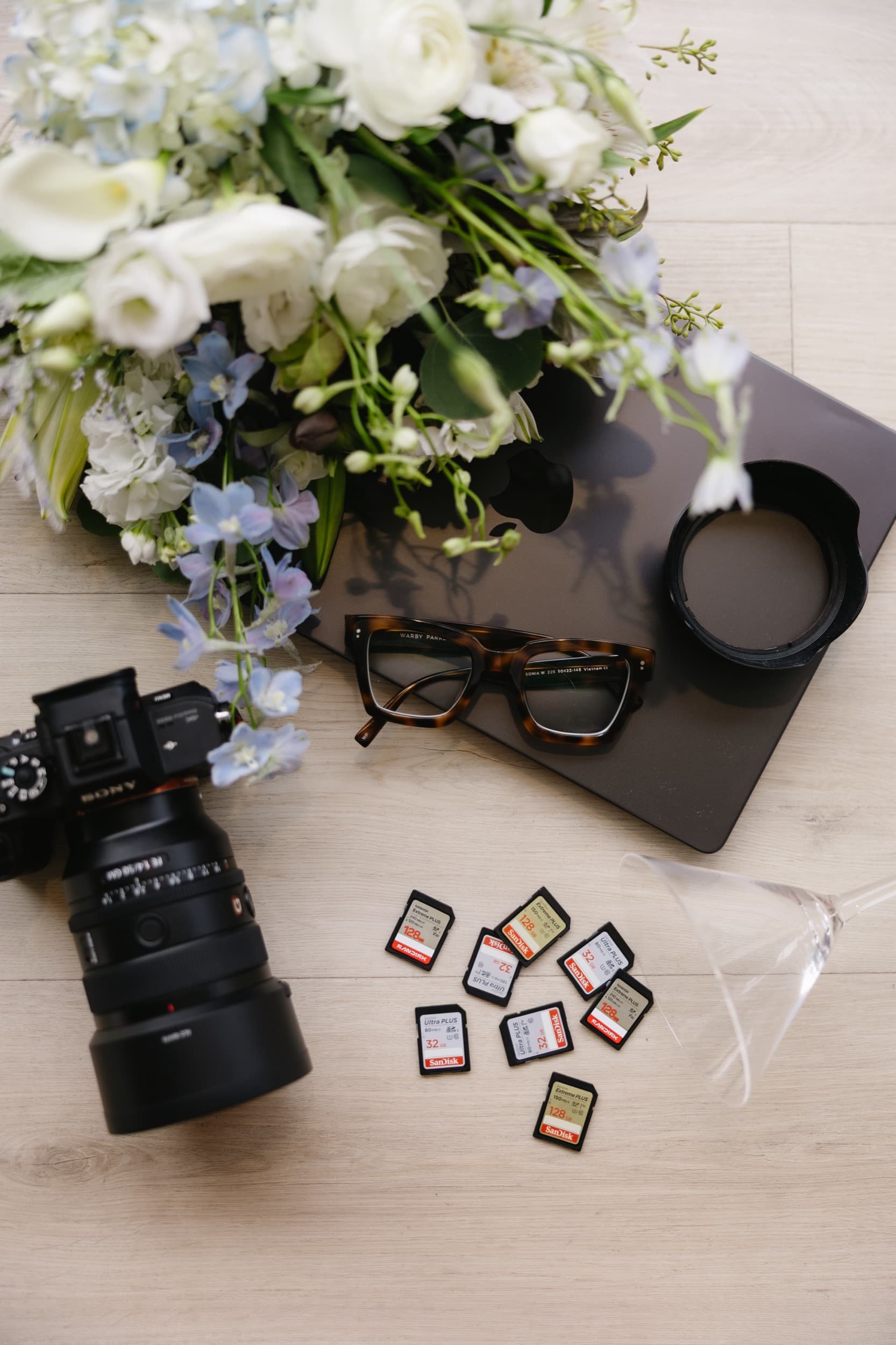 Flat lay of a camera, glasses, memory cards, and a white and blue floral bouquet on wood flooring.