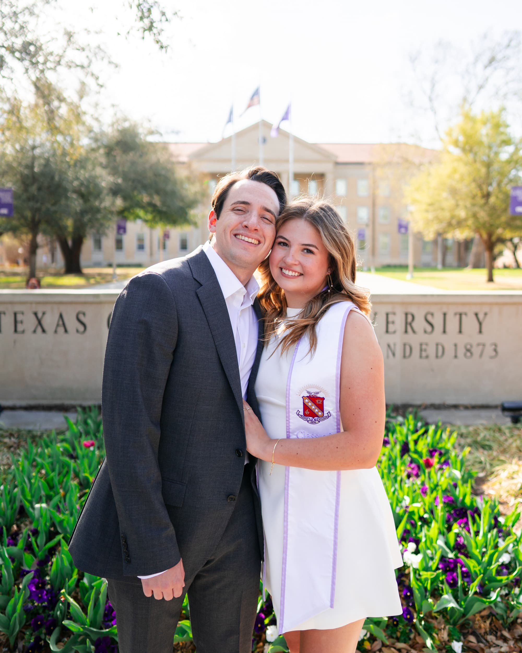 Couple embracing warmly by a university sign, surrounded by purple and white pansies.