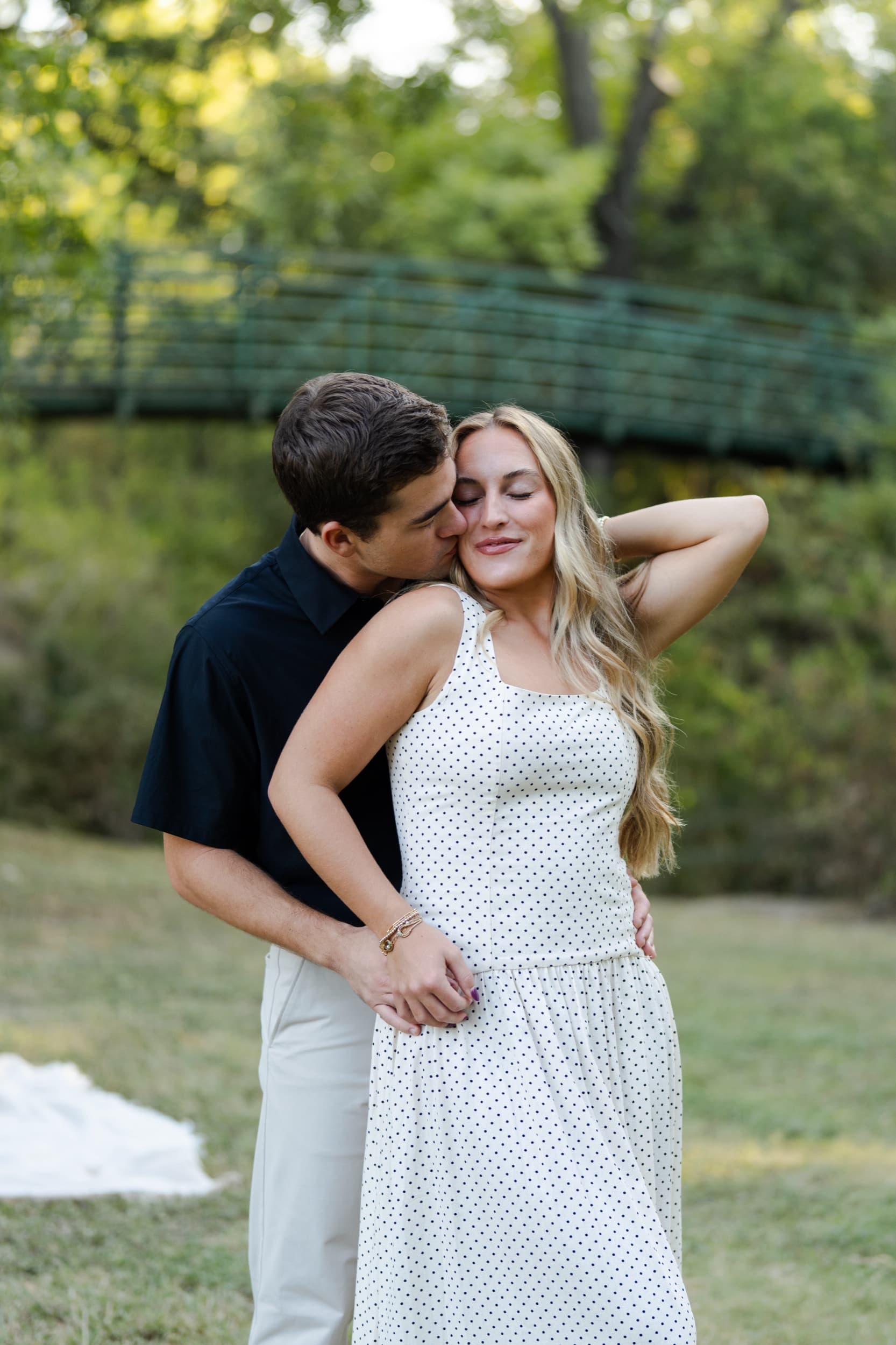 Man kissing his partner's cheek as she smiles with her eyes closed, a green footbridge in the background.