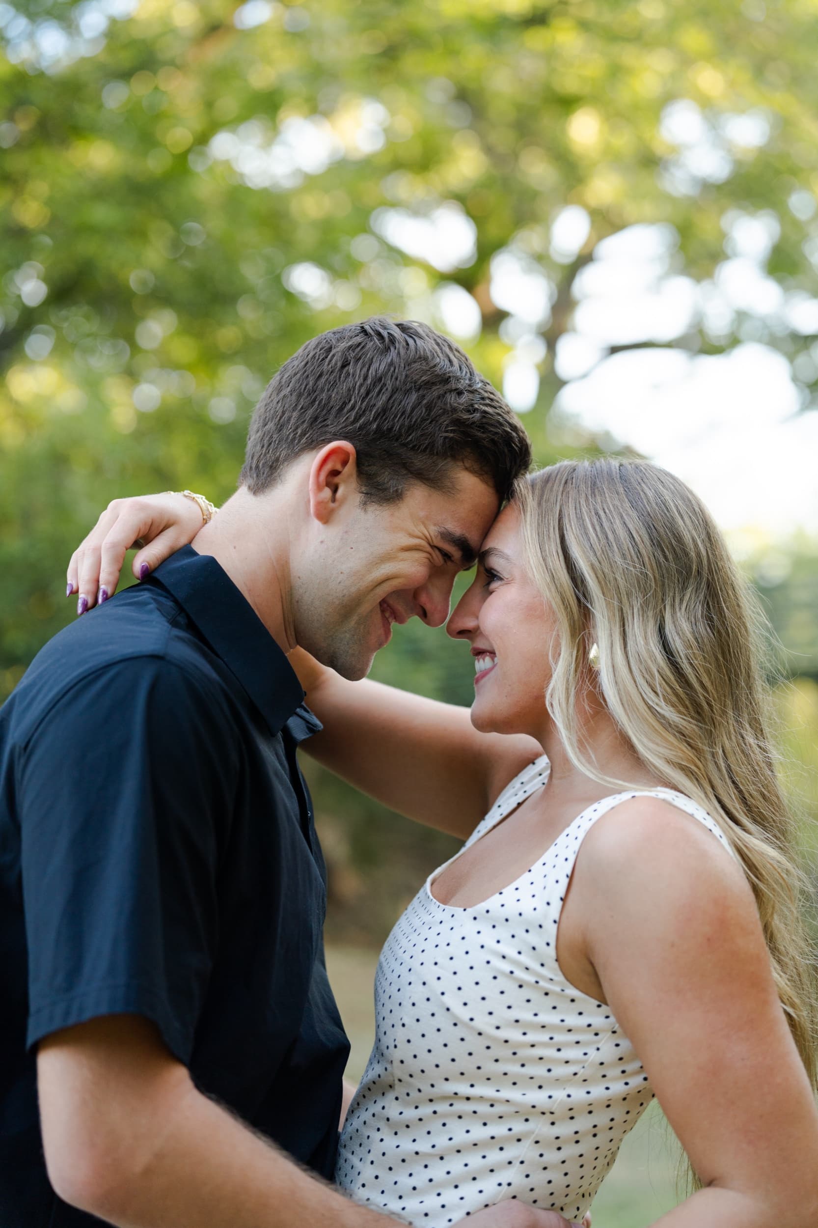 Couple laughing forehead to forehead, her arm around his shoulders, in a sunlit park.