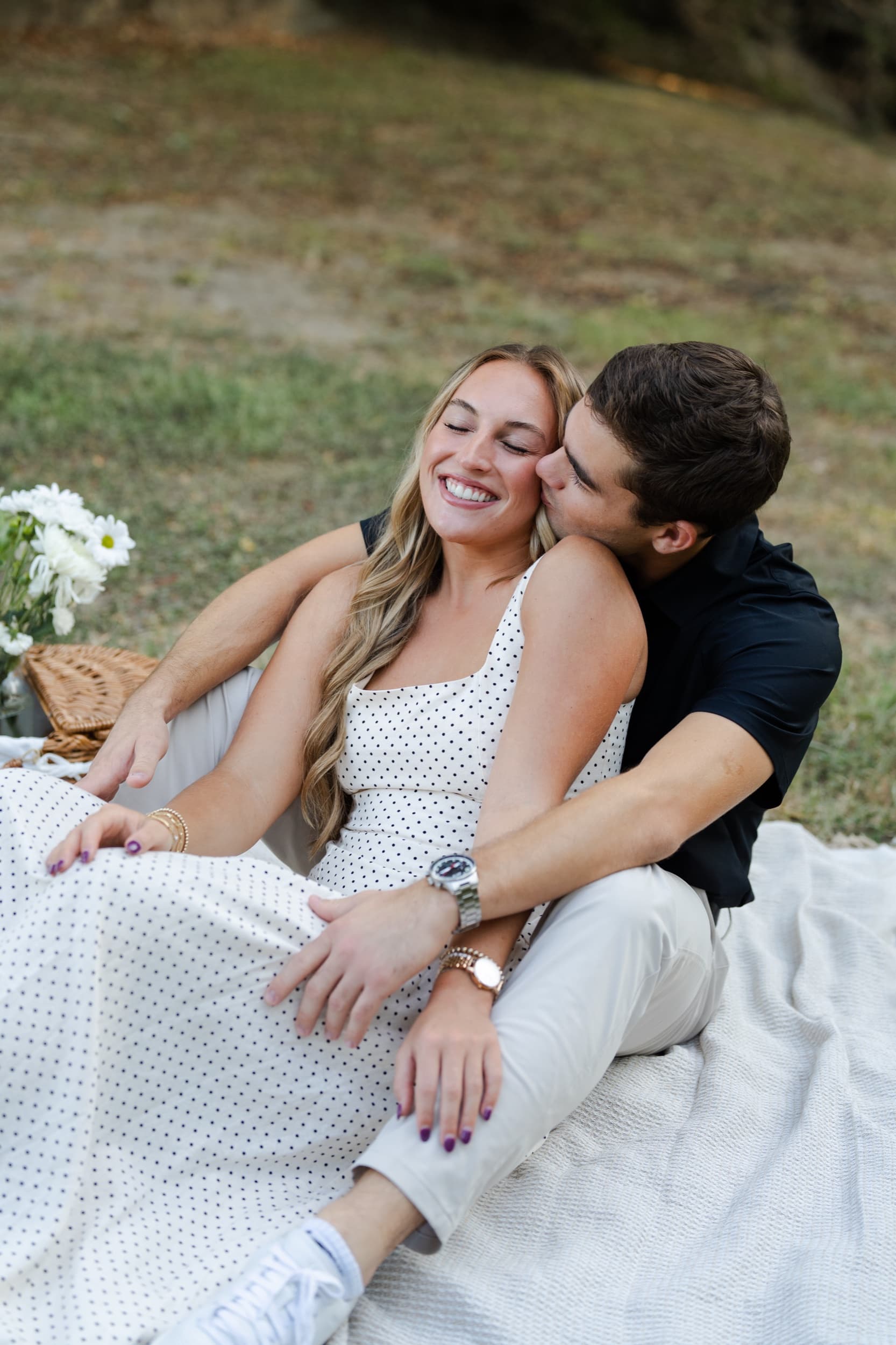 Couple laughing together on a picnic blanket, him kissing her cheek beside a basket of white flowers.