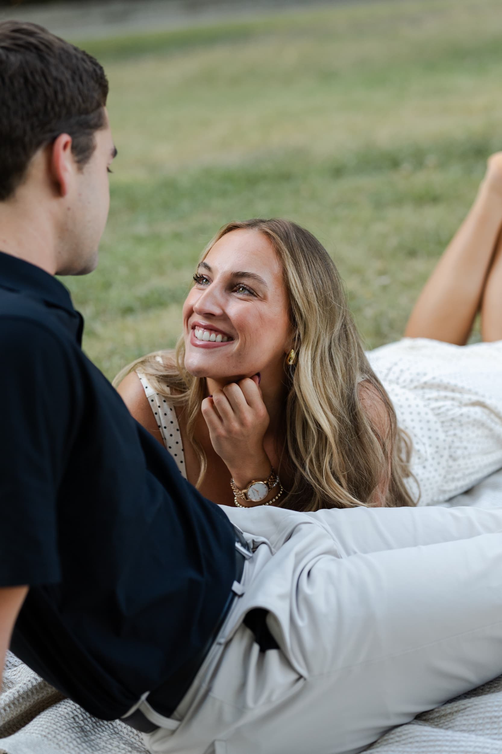 Woman resting her chin on her hand, smiling at her partner while lying on a picnic blanket in a grassy field.
