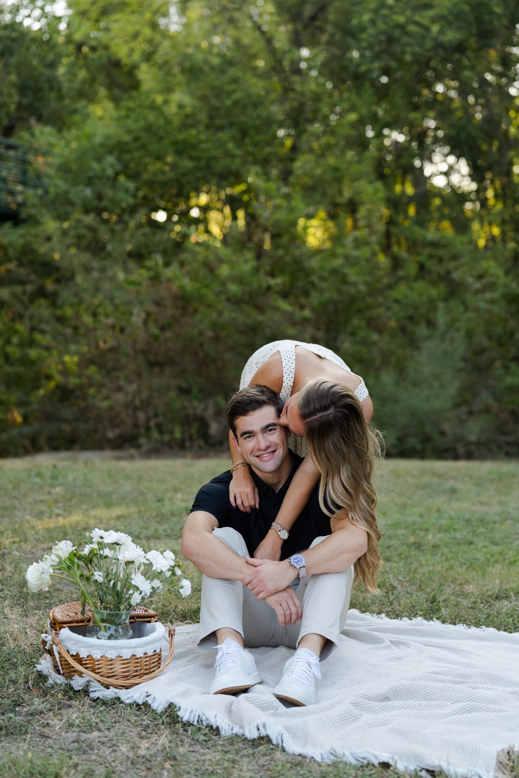Woman leaning down to kiss her partner's cheek as he smiles, sitting on a picnic blanket beside a basket of flowers.