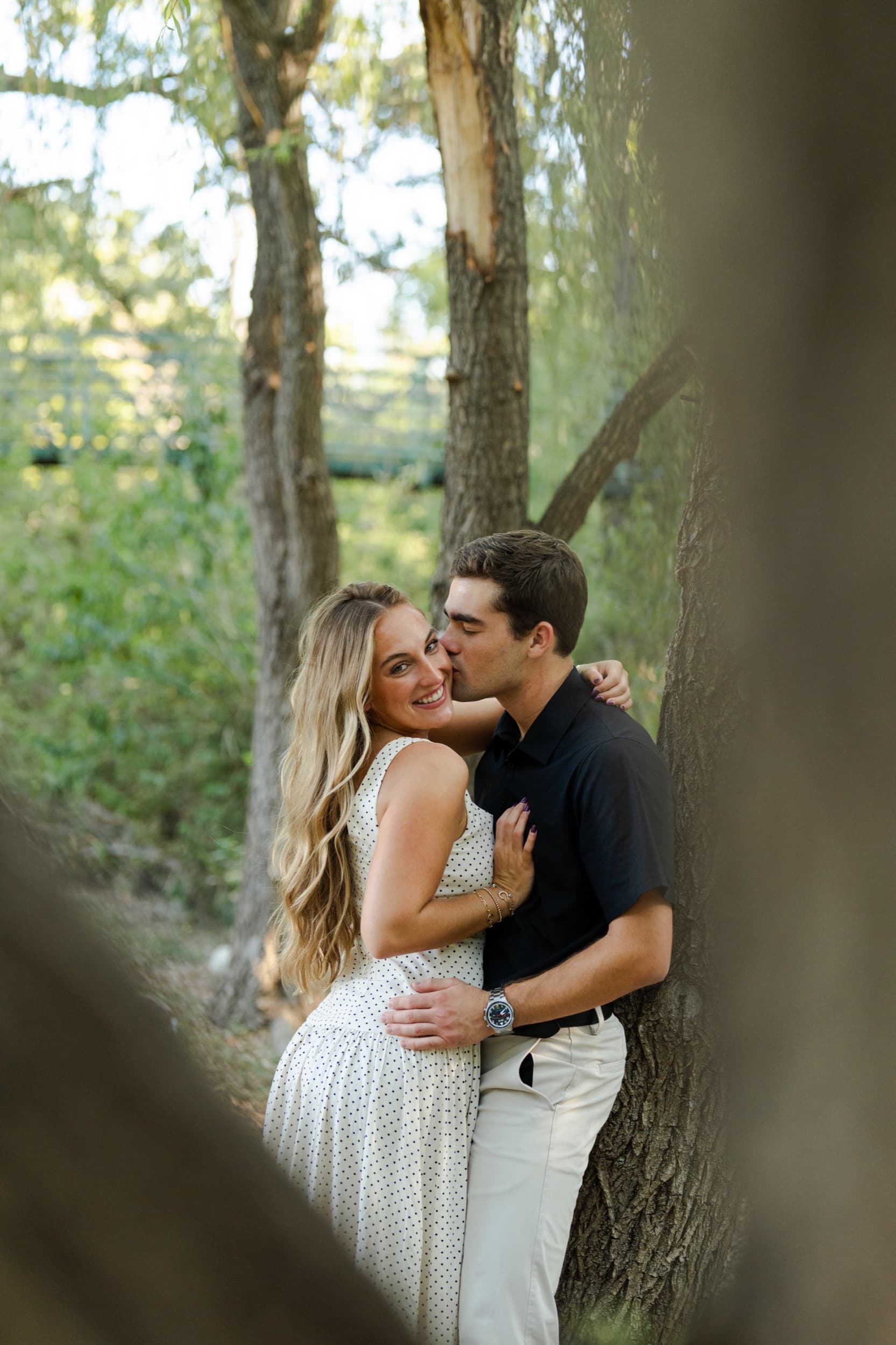Man kissing his smiling partner's cheek while she looks at the camera, framed between trees.