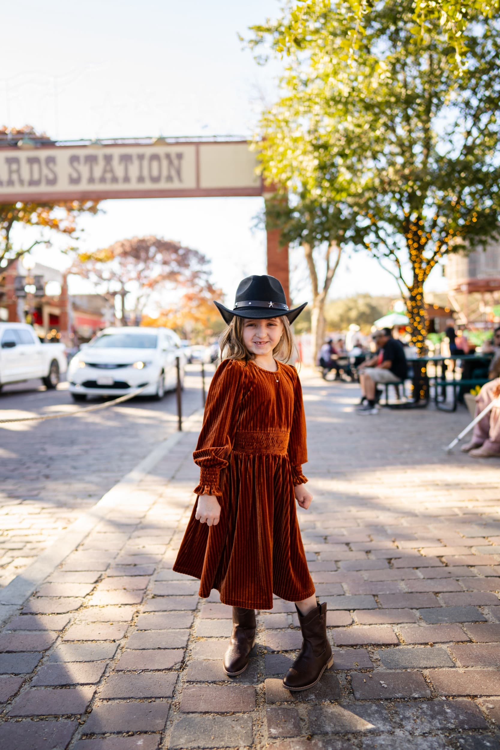 Little girl in a velvet dress and cowboy hat standing on a cobblestone street.