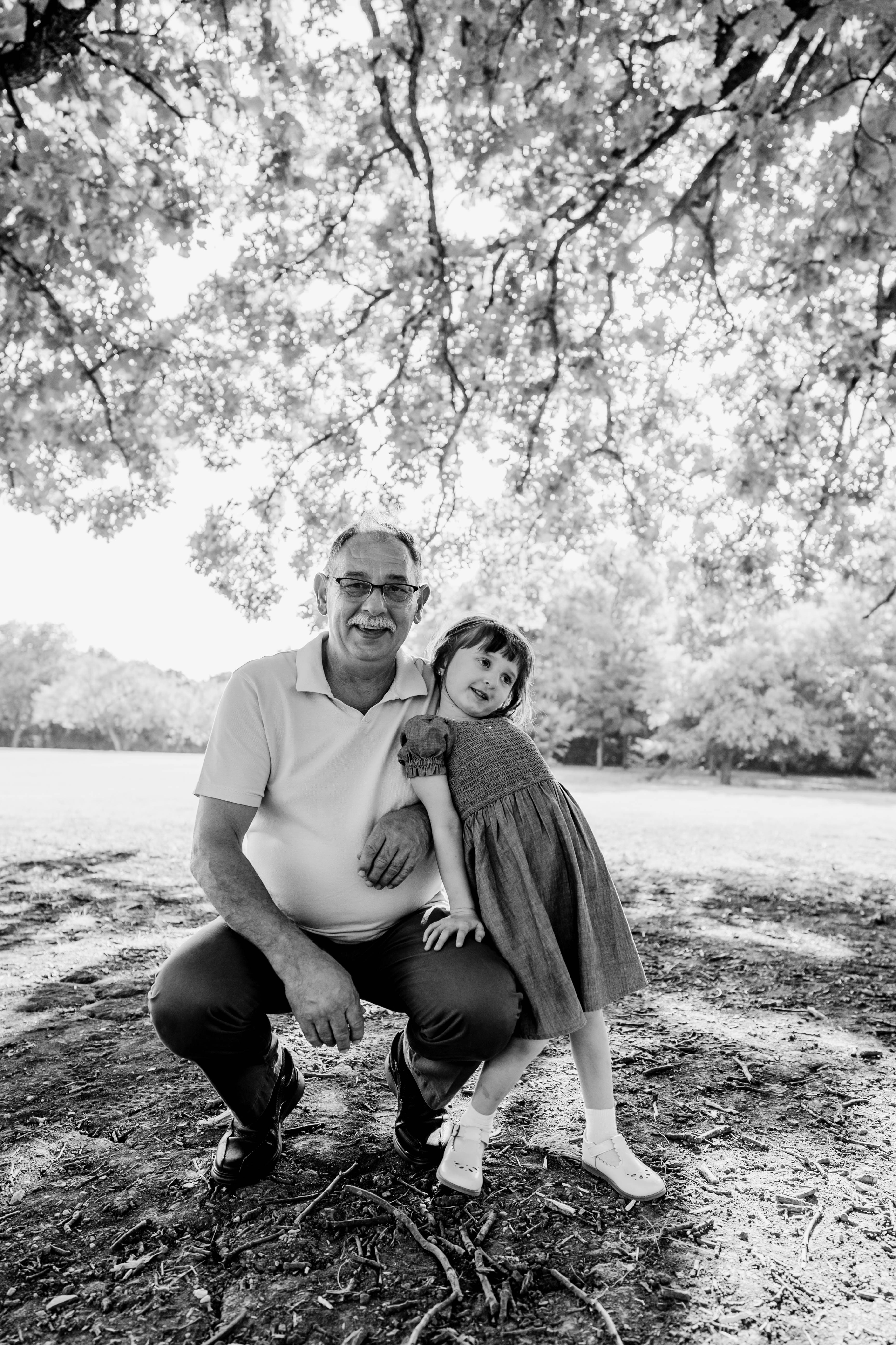 Grandfather kneeling beside his granddaughter under a shaded tree, both smiling together.