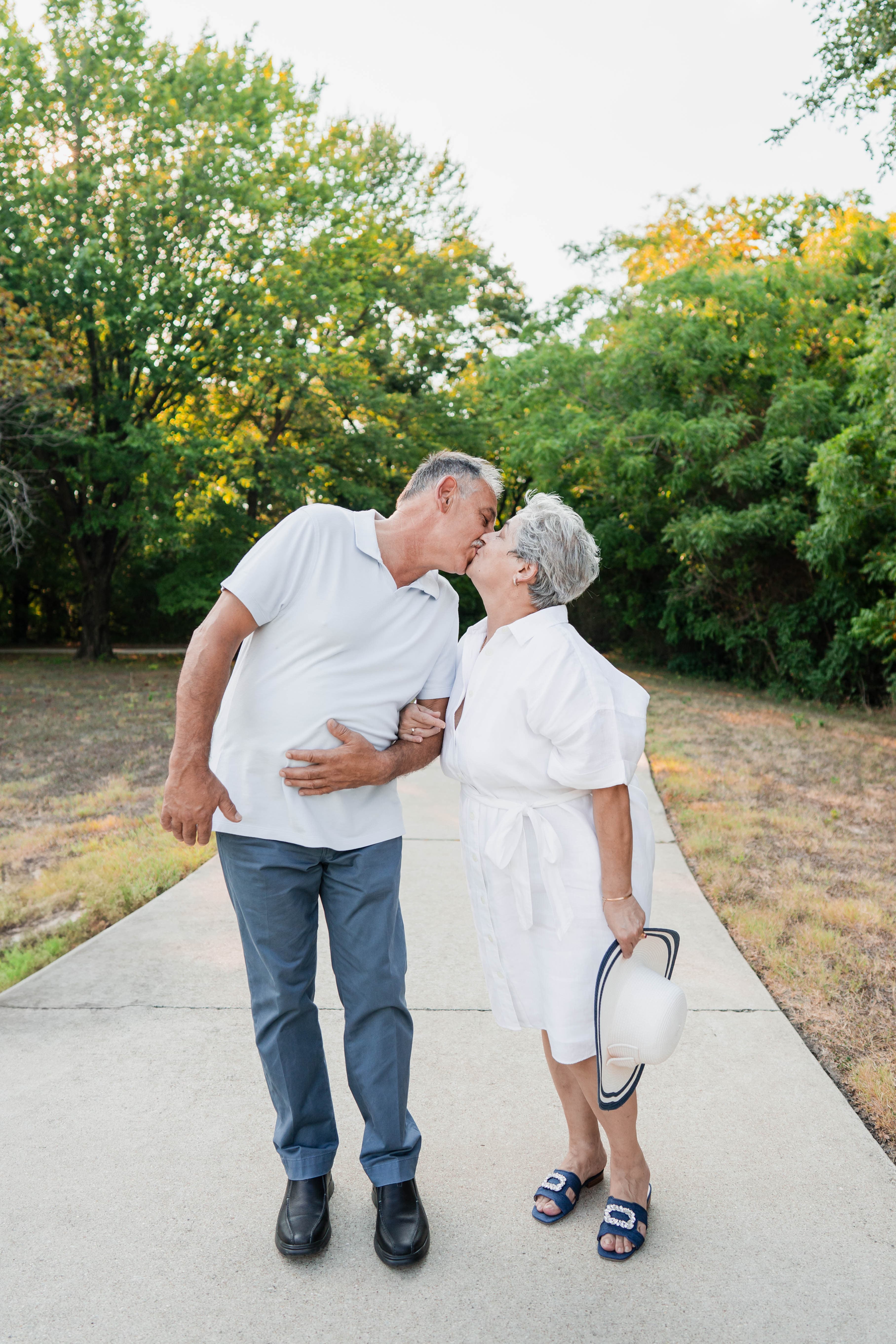 Grandparents sharing a kiss on a path, one holding a sun hat.