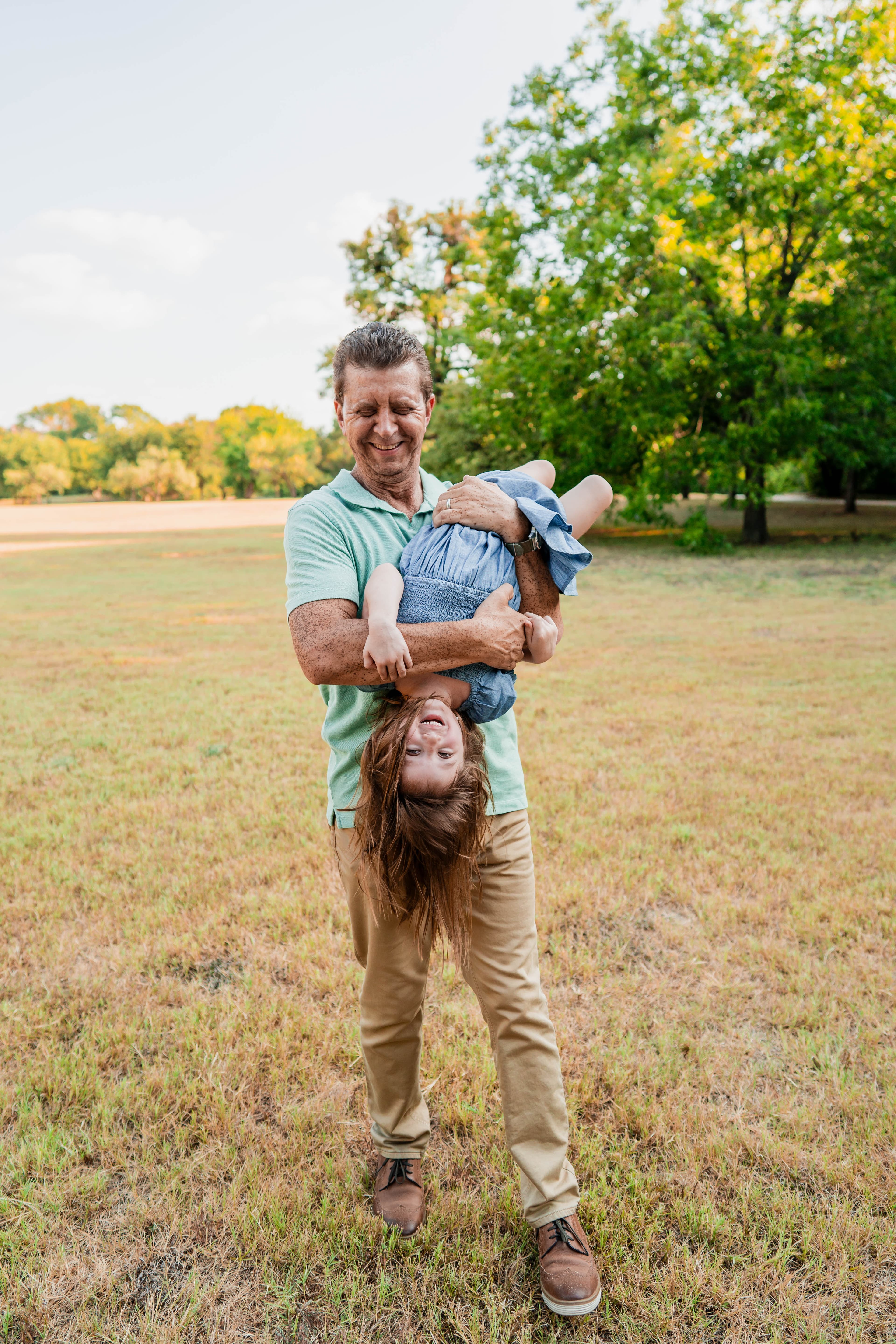 Grandfather laughing while holding his granddaughter upside down in an open field.