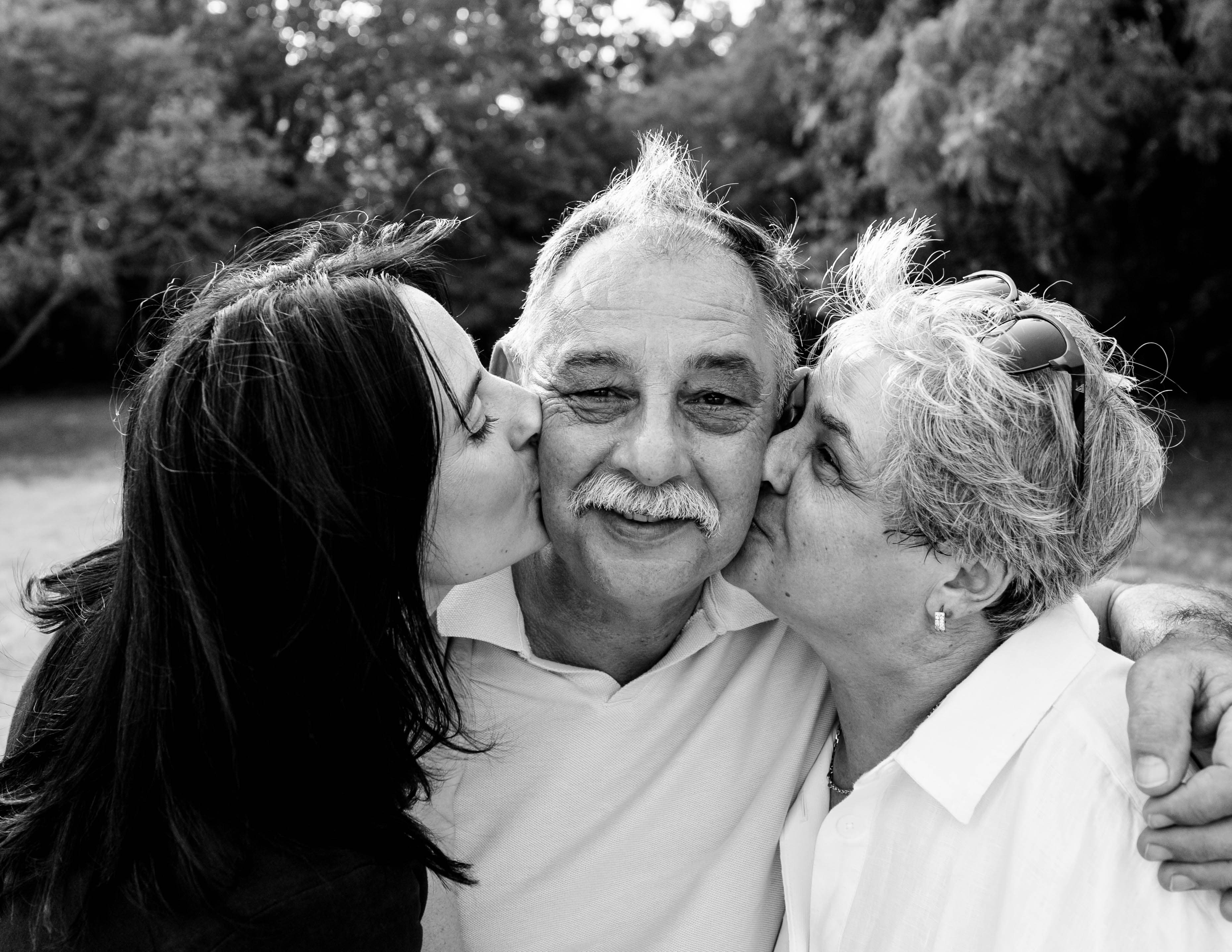 Grandfather smiling as two family members kiss him on each cheek.
