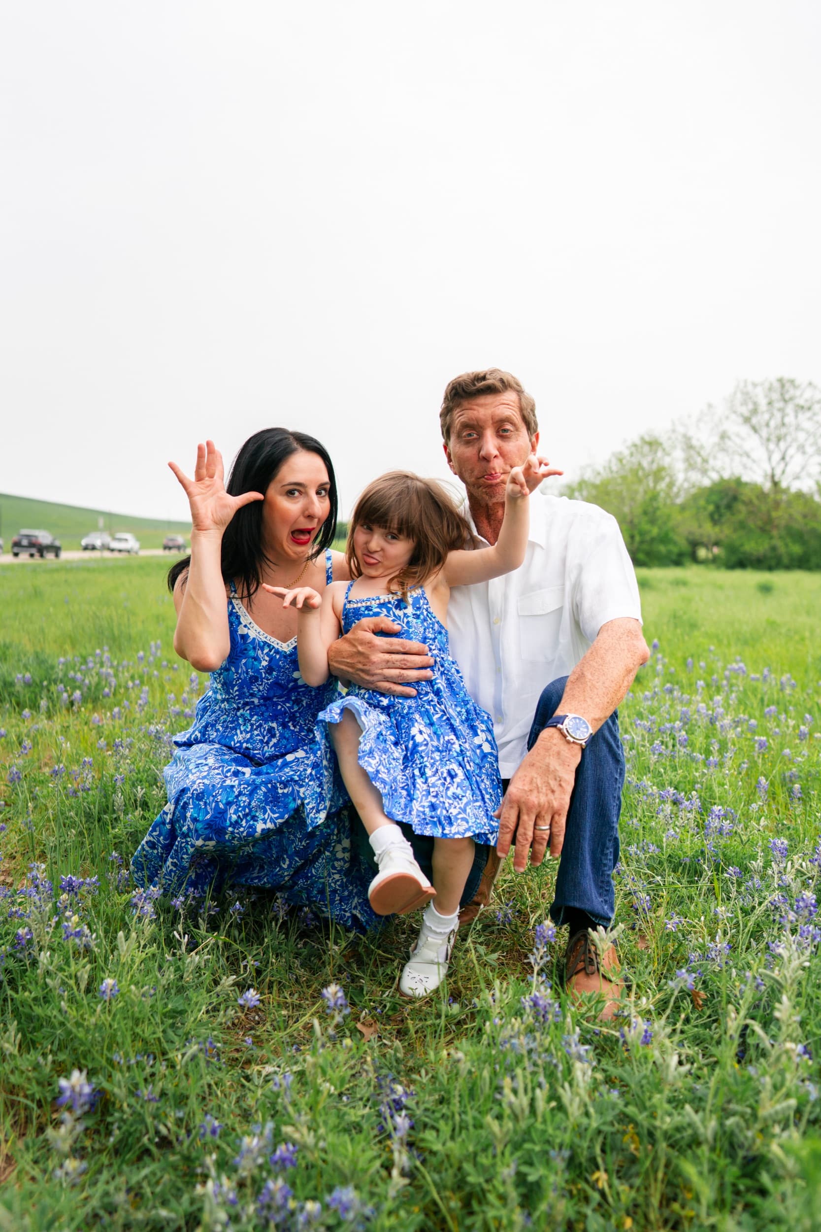 Family in matching blue outfits making playful silly faces in a bluebonnet field.