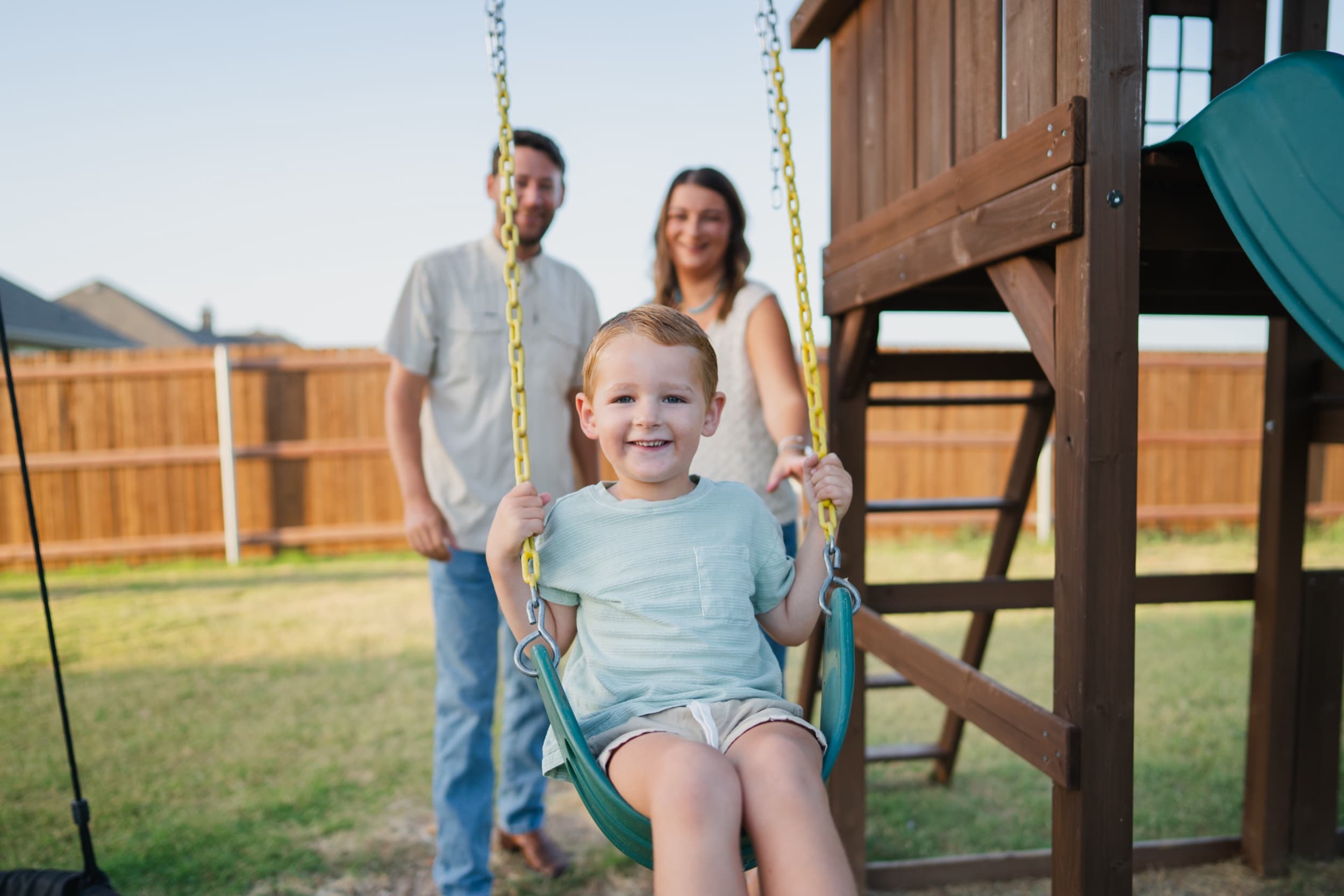 Little boy laughing on a swing while his parents look on from behind at a backyard play set.