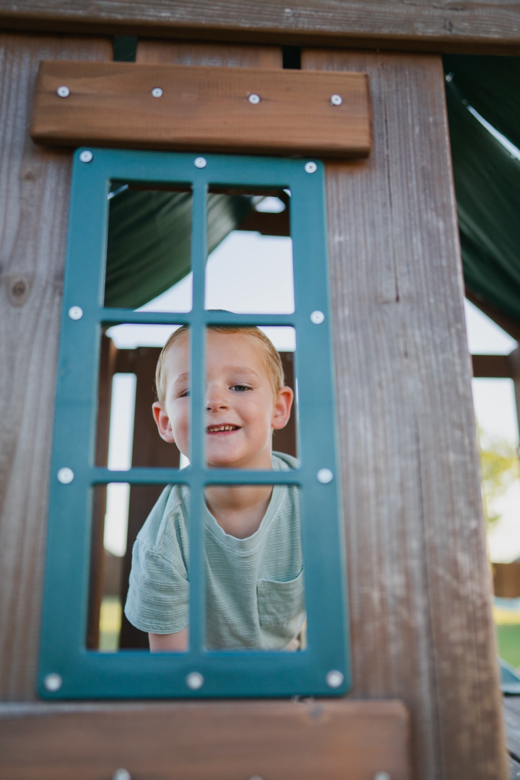 Little boy peeking through a play set's window frame with a big smile.