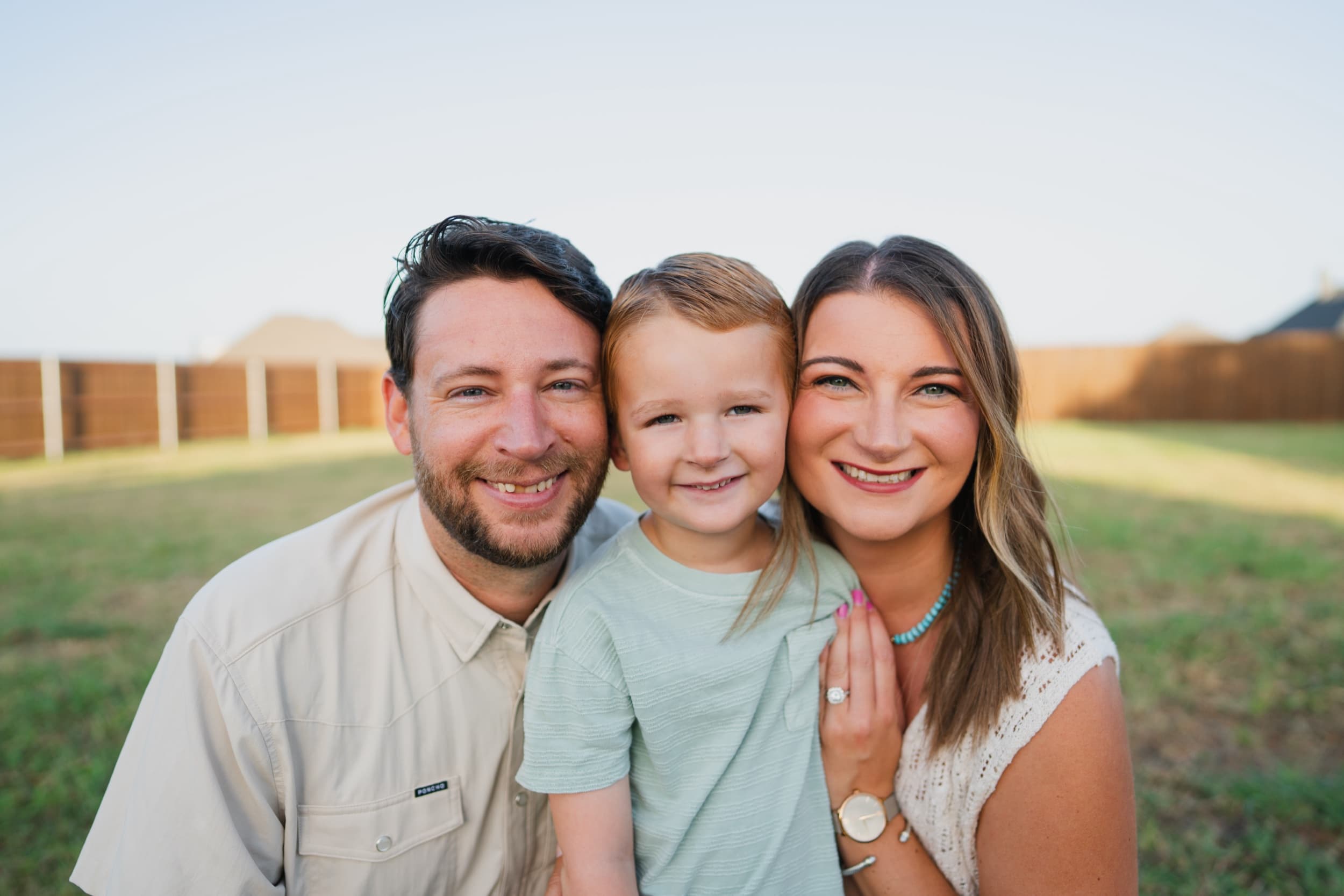 Mom and dad smiling close together with their son between them in a backyard.