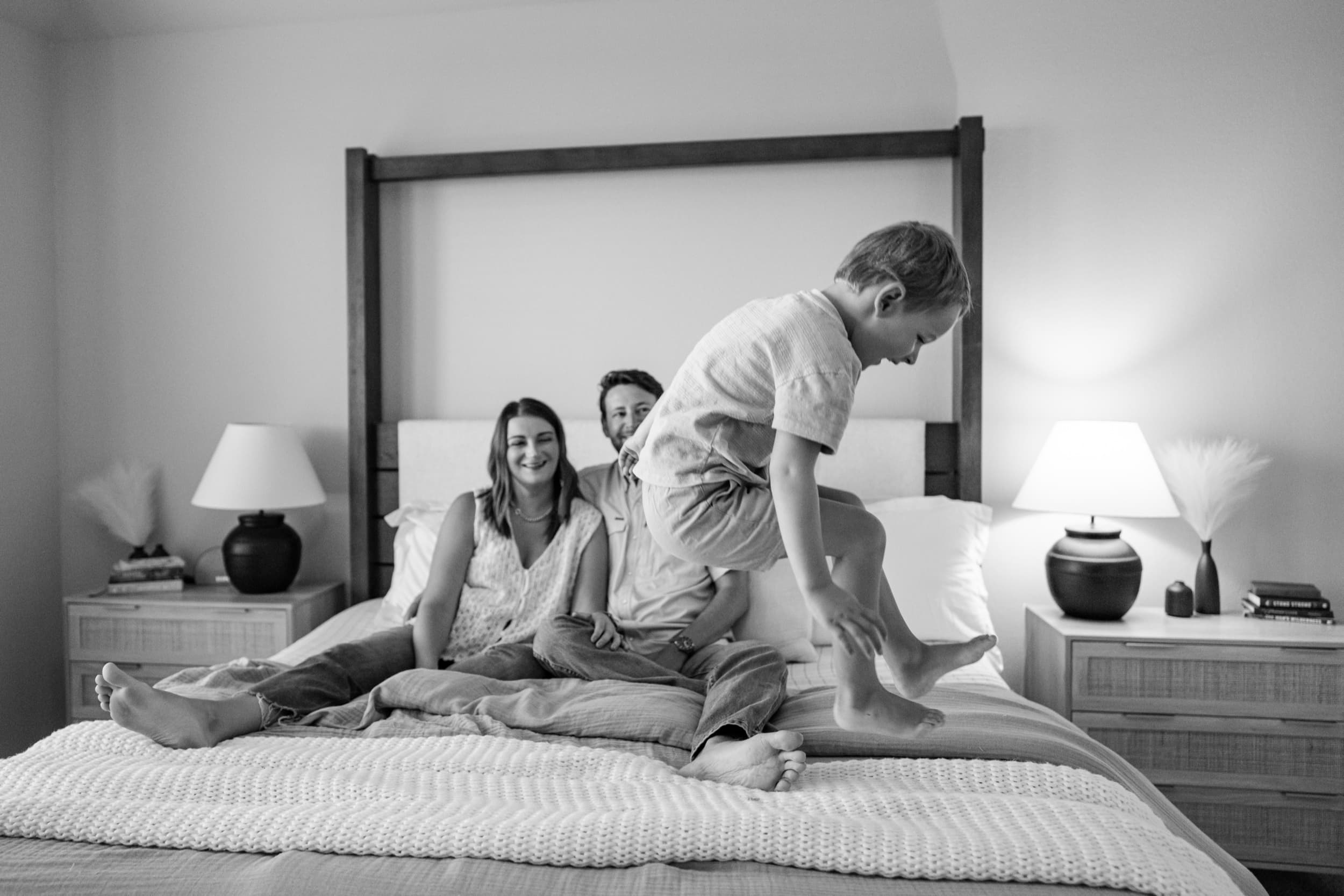 Black and white photo of a boy leaping off the bed while his parents laugh in the background.