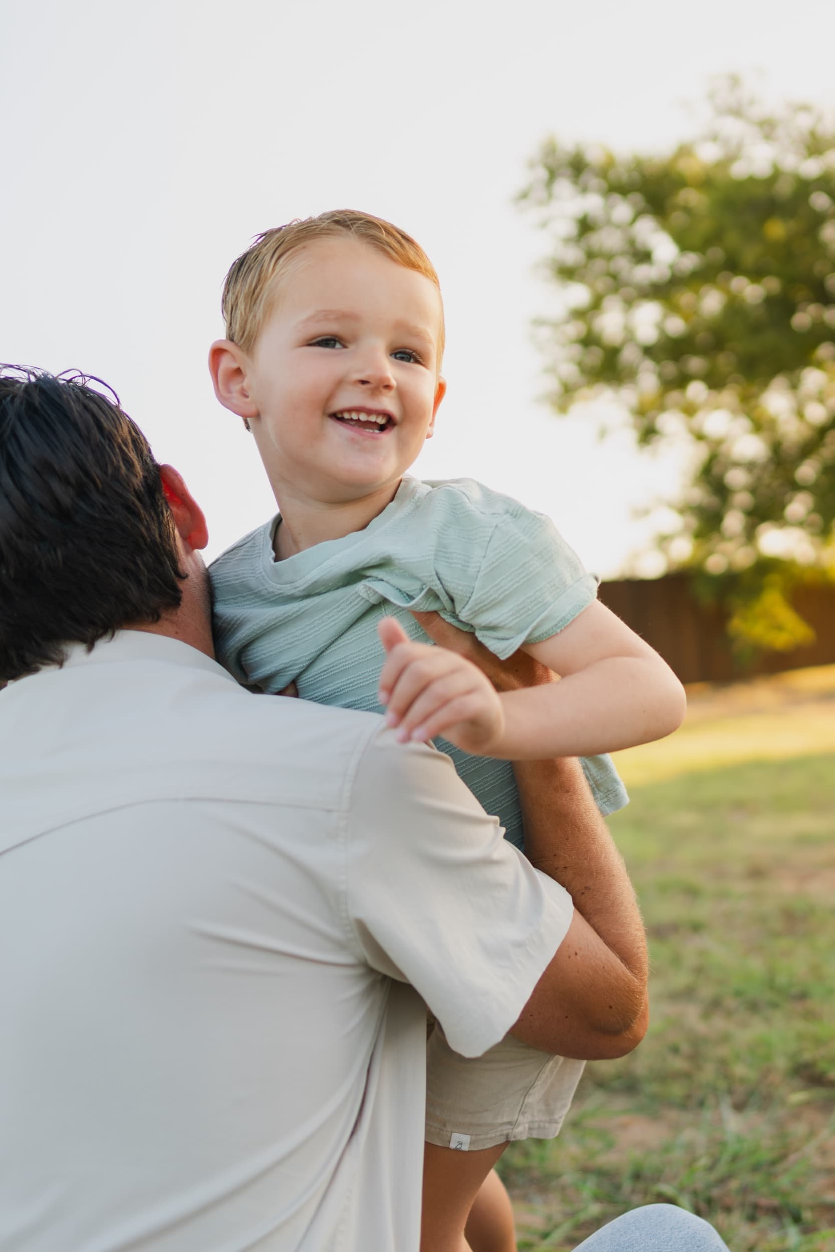 Dad carrying his laughing son on his hip outdoors at golden hour.