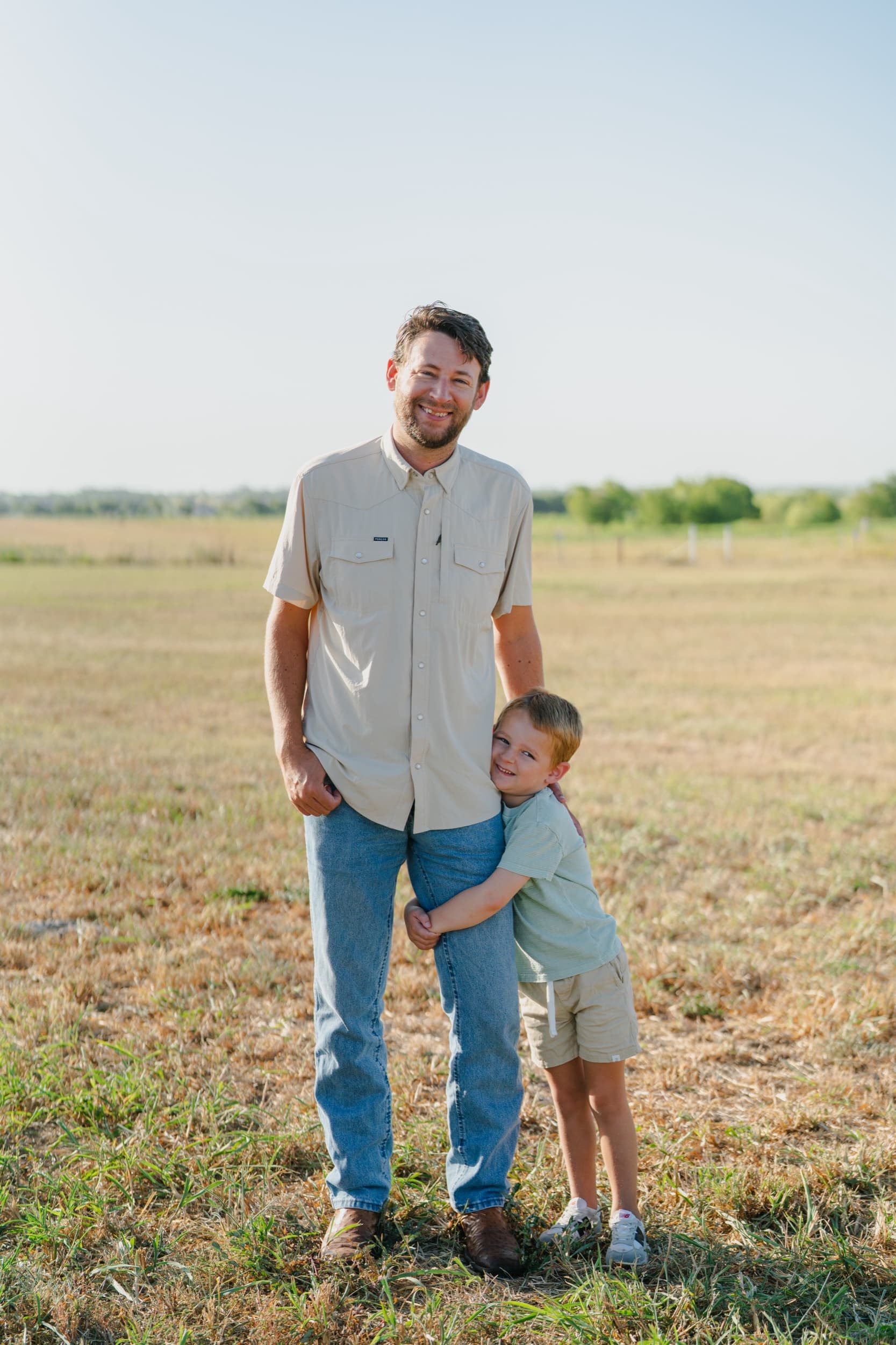 Little boy hugging his dad's leg while both smile in an open field.