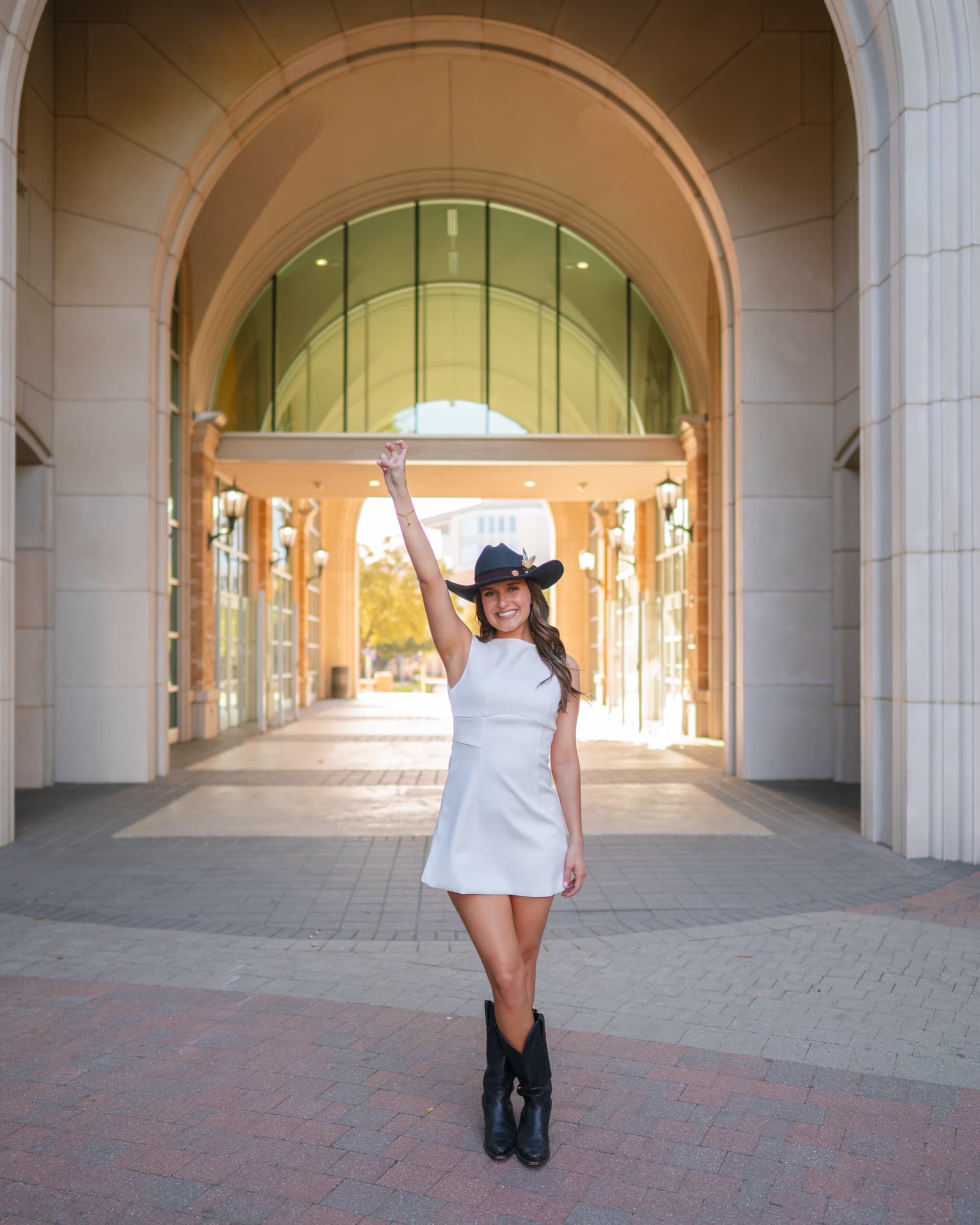 Senior in a white dress and cowboy hat raising her arm under a sunlit archway.