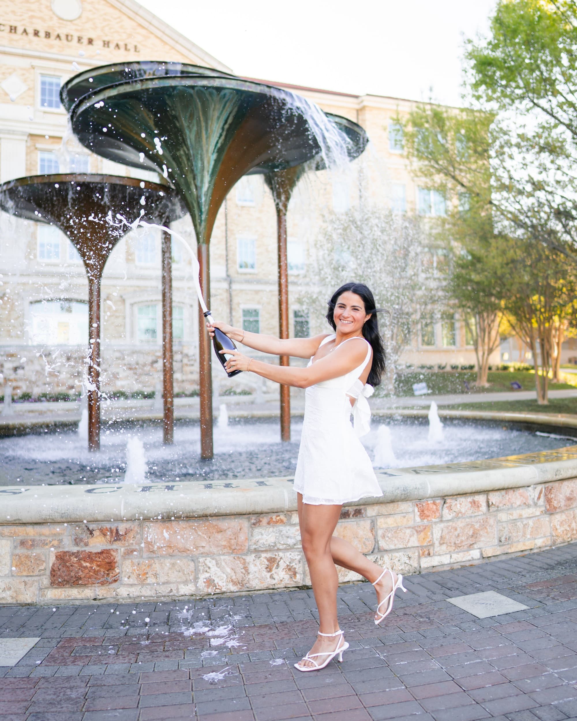 Senior dancing joyfully in a white dress while popping champagne beside a fountain.