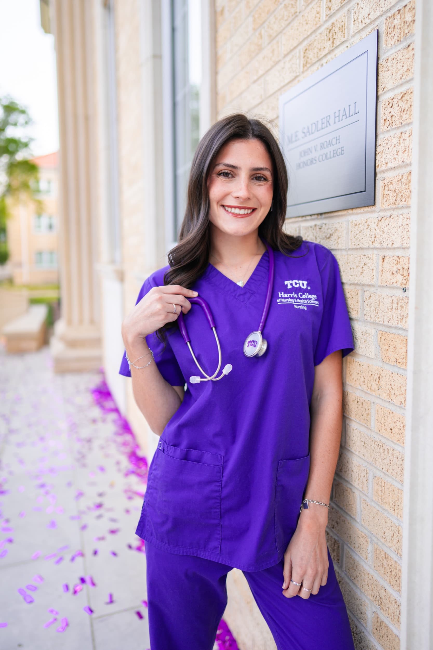 Nursing graduate in purple scrubs holding a stethoscope, smiling beside a brick building.