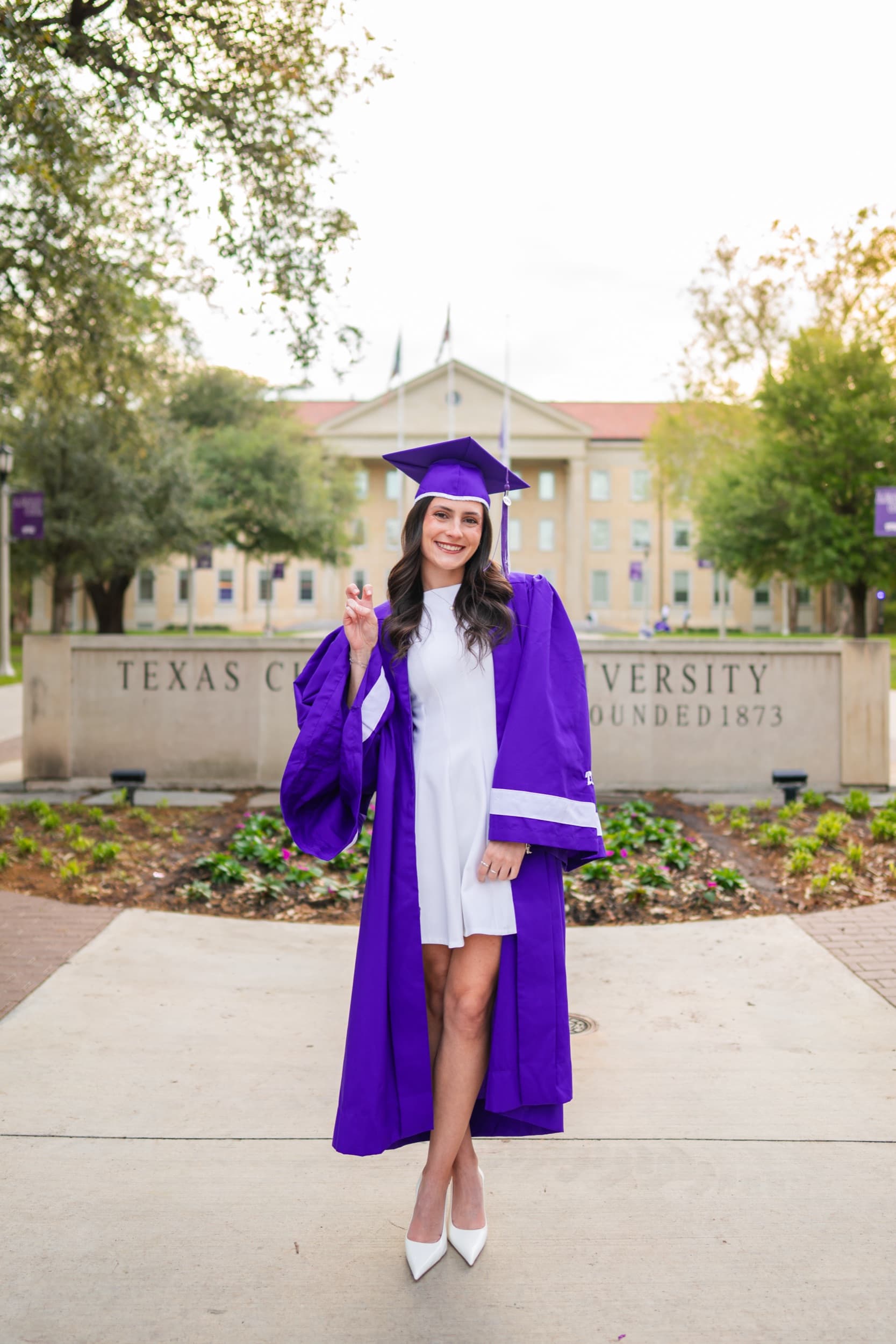 Graduate in cap and gown crossing fingers playfully in front of a university sign.