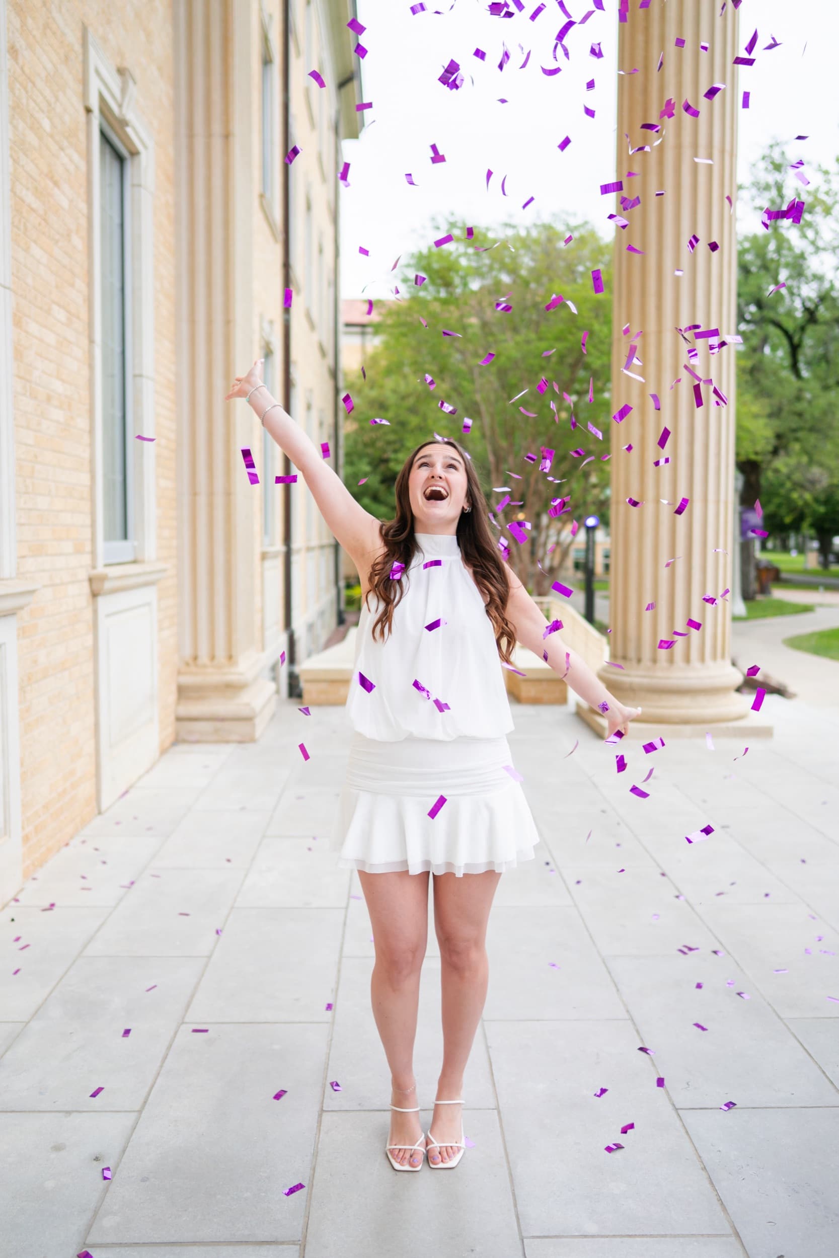 Senior throwing purple confetti into the air, laughing joyfully beside stone columns.