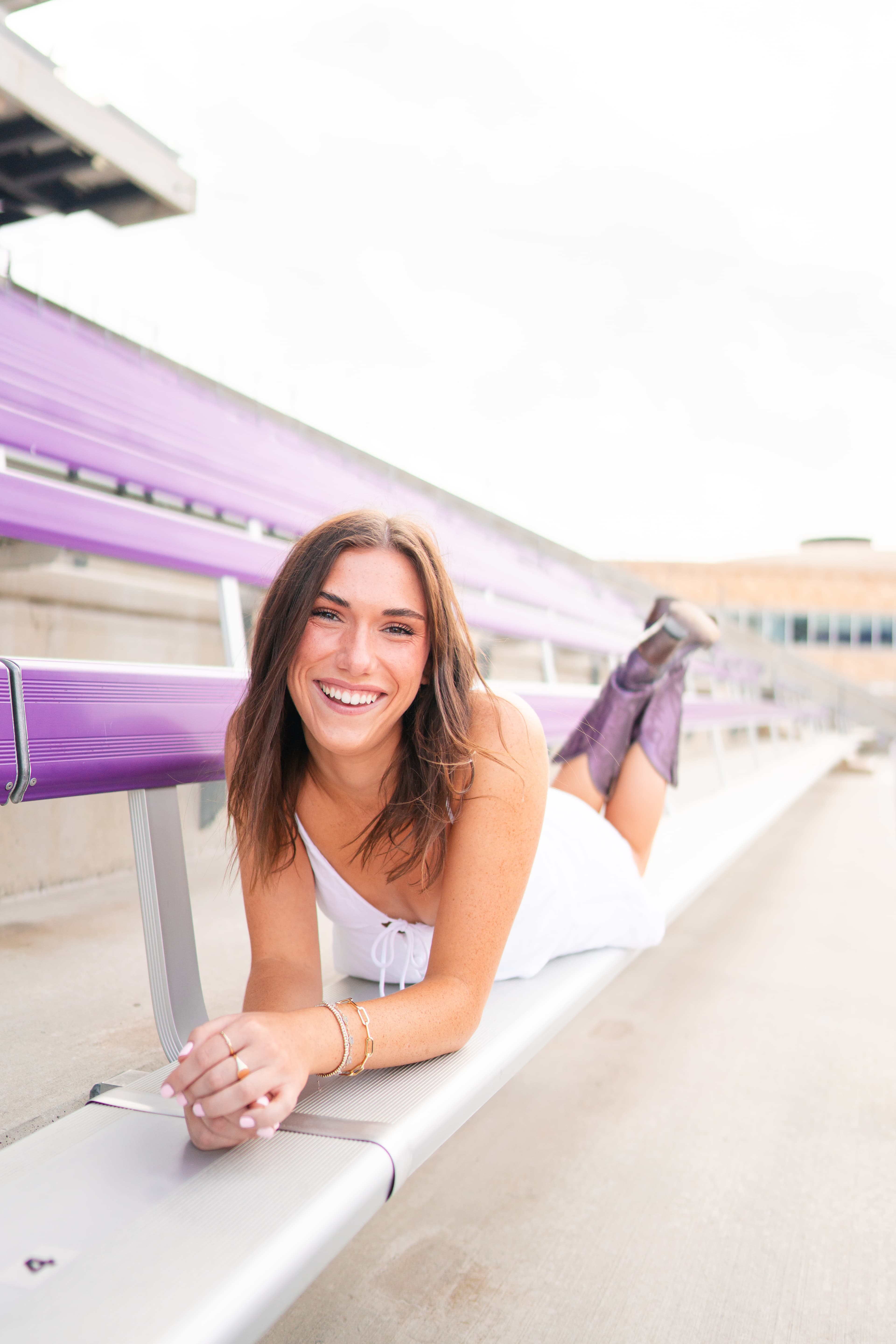 Senior lying on purple stadium bleachers in a white dress, smiling with hands folded and heels crossed in the air.