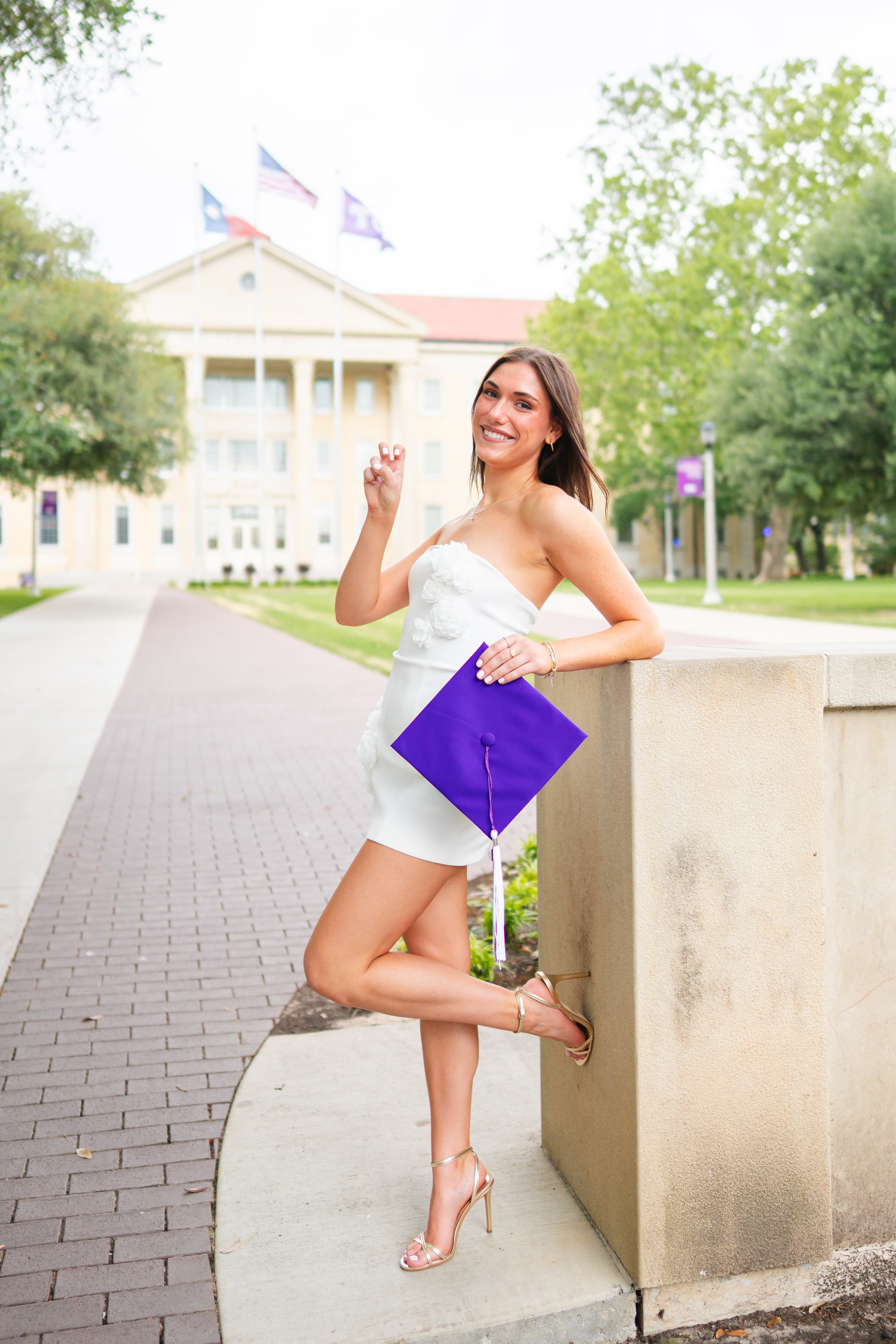 Graduate in a white dress holding a purple graduation cap, posing against a stone wall in front of a university building.