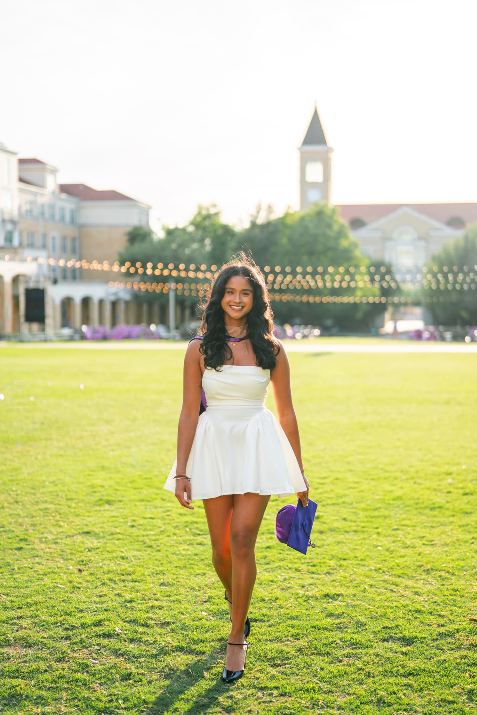 Senior walking across a sunlit lawn, holding her graduation cap near a clock tower.