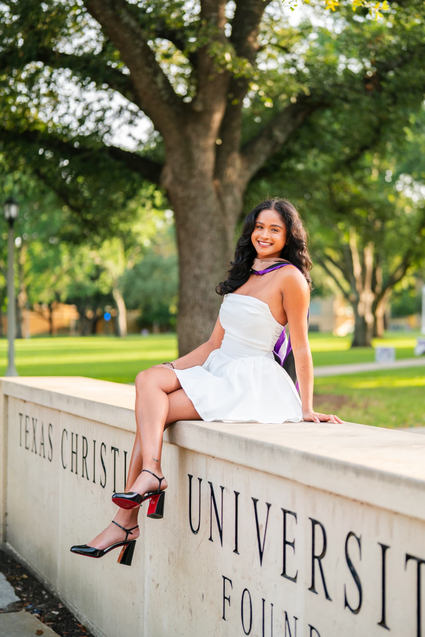 Senior in a white dress sitting on a university sign wall under shady trees.