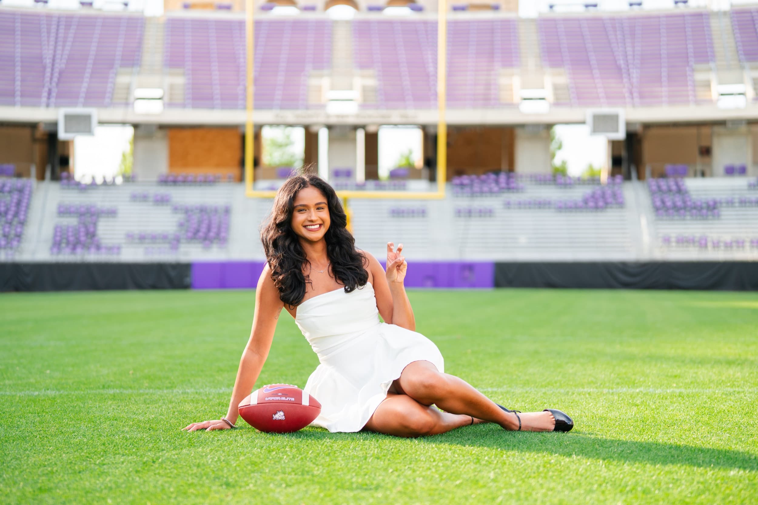 Senior in a white dress sitting on the football field beside a football, smiling brightly.