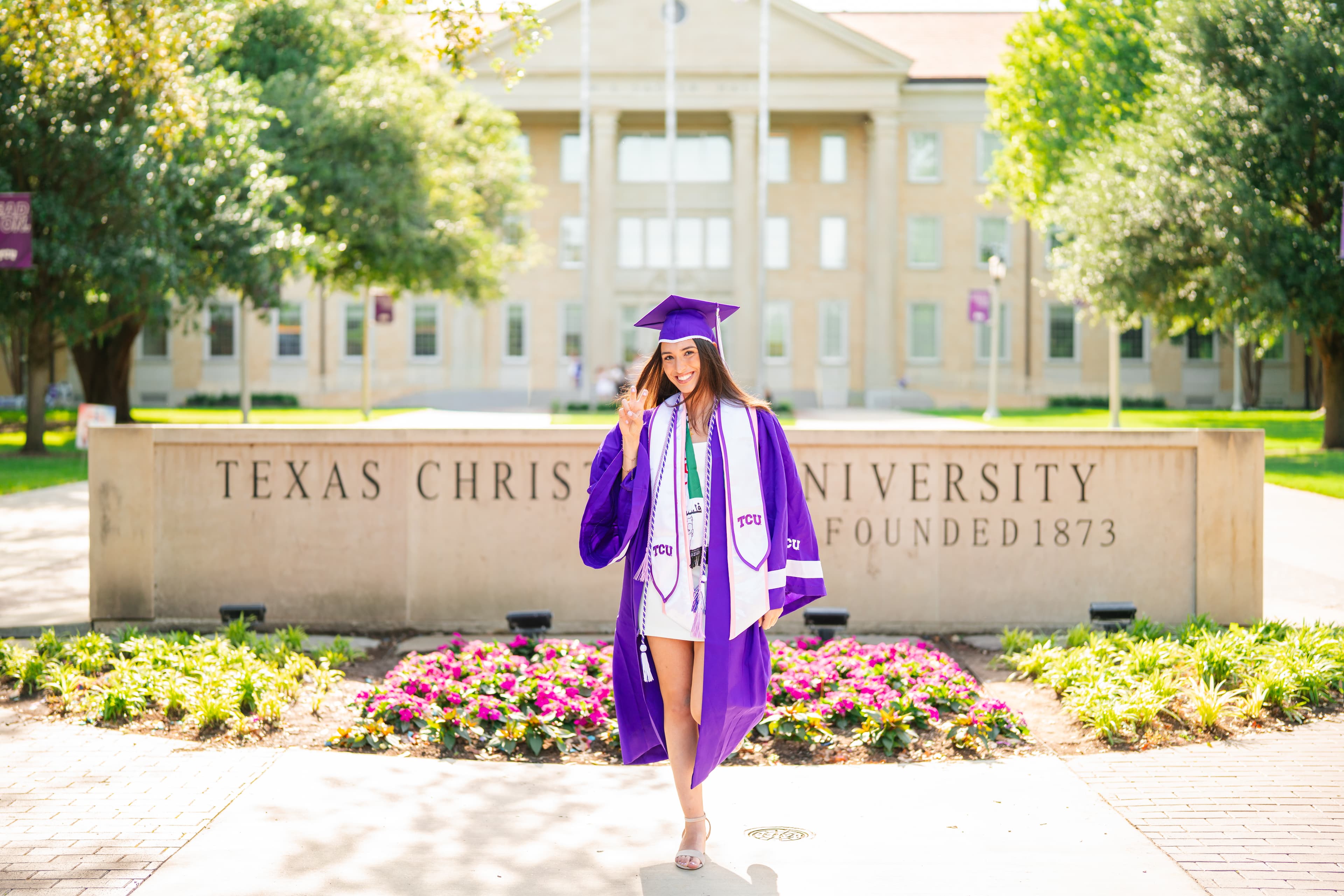 Graduate in a cap and gown with honor stoles giving a peace sign in front of a university sign.