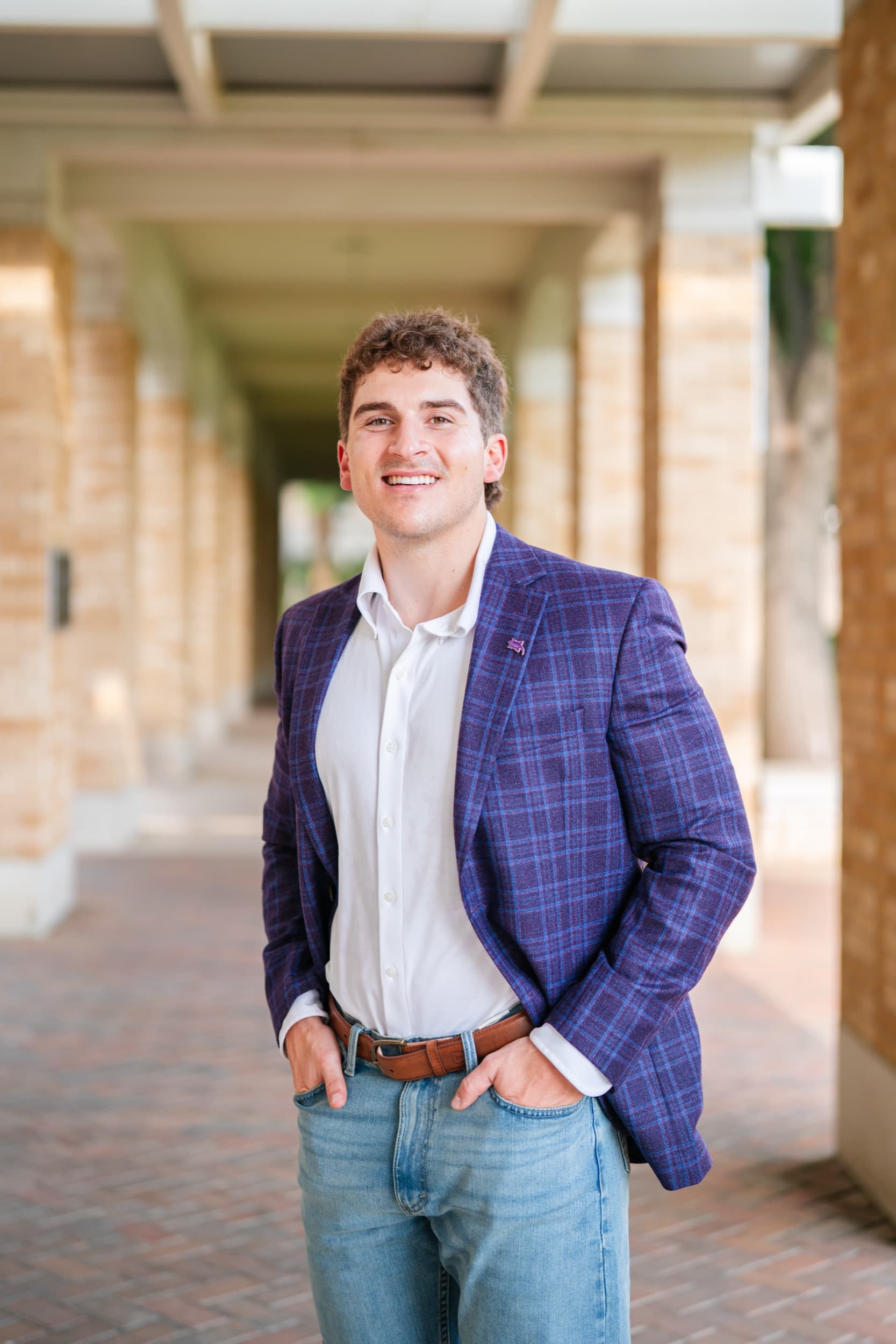 Senior in a plaid blazer smiling confidently under a sunlit stone colonnade.