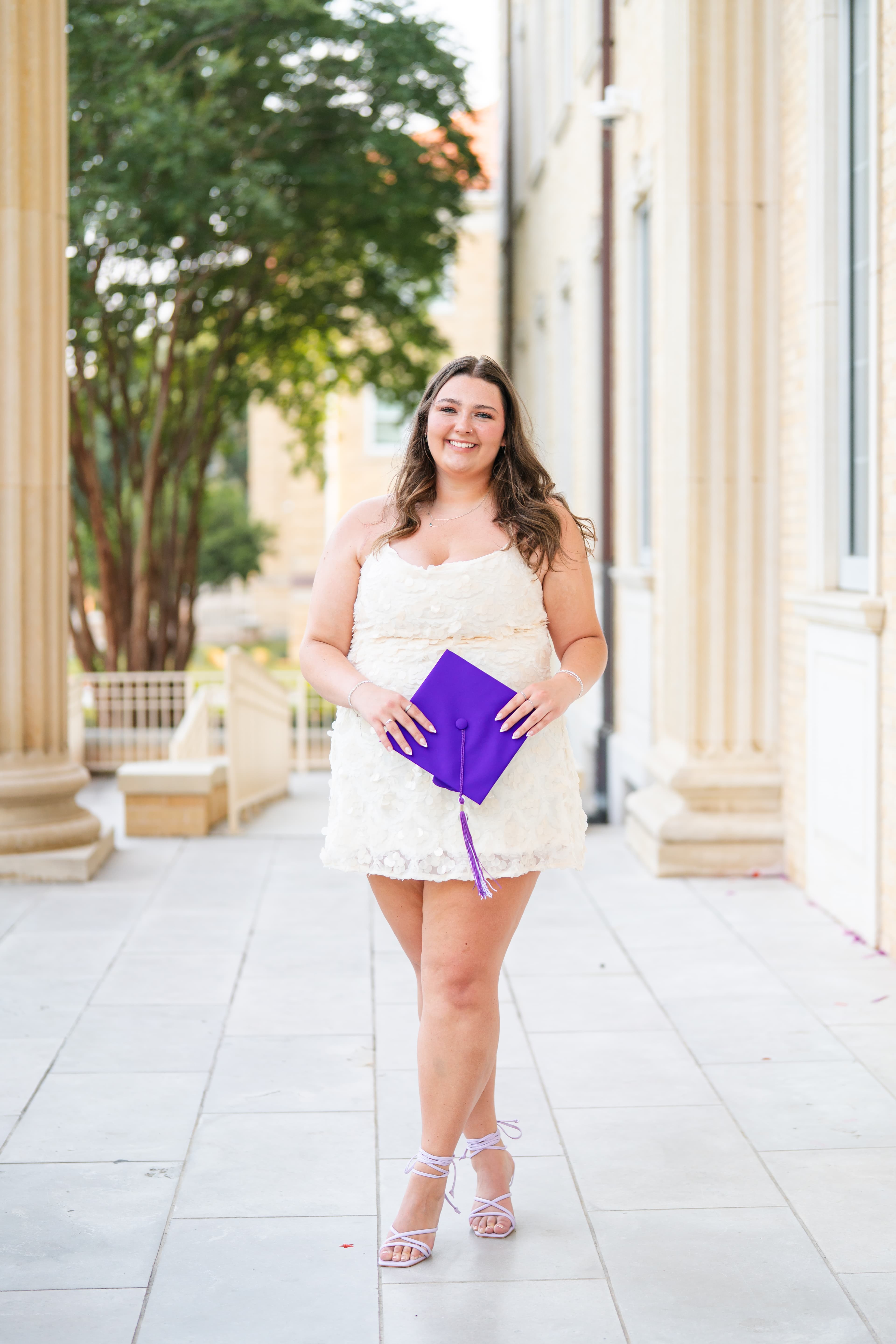 Graduate in a white dress holding her purple graduation cap, smiling on a sunlit university walkway.