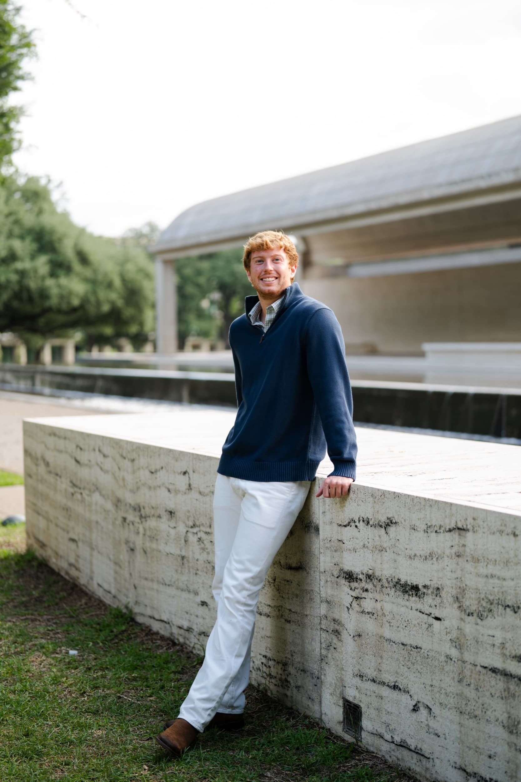 Senior leaning against a concrete wall near a modern fountain, smiling casually outdoors.