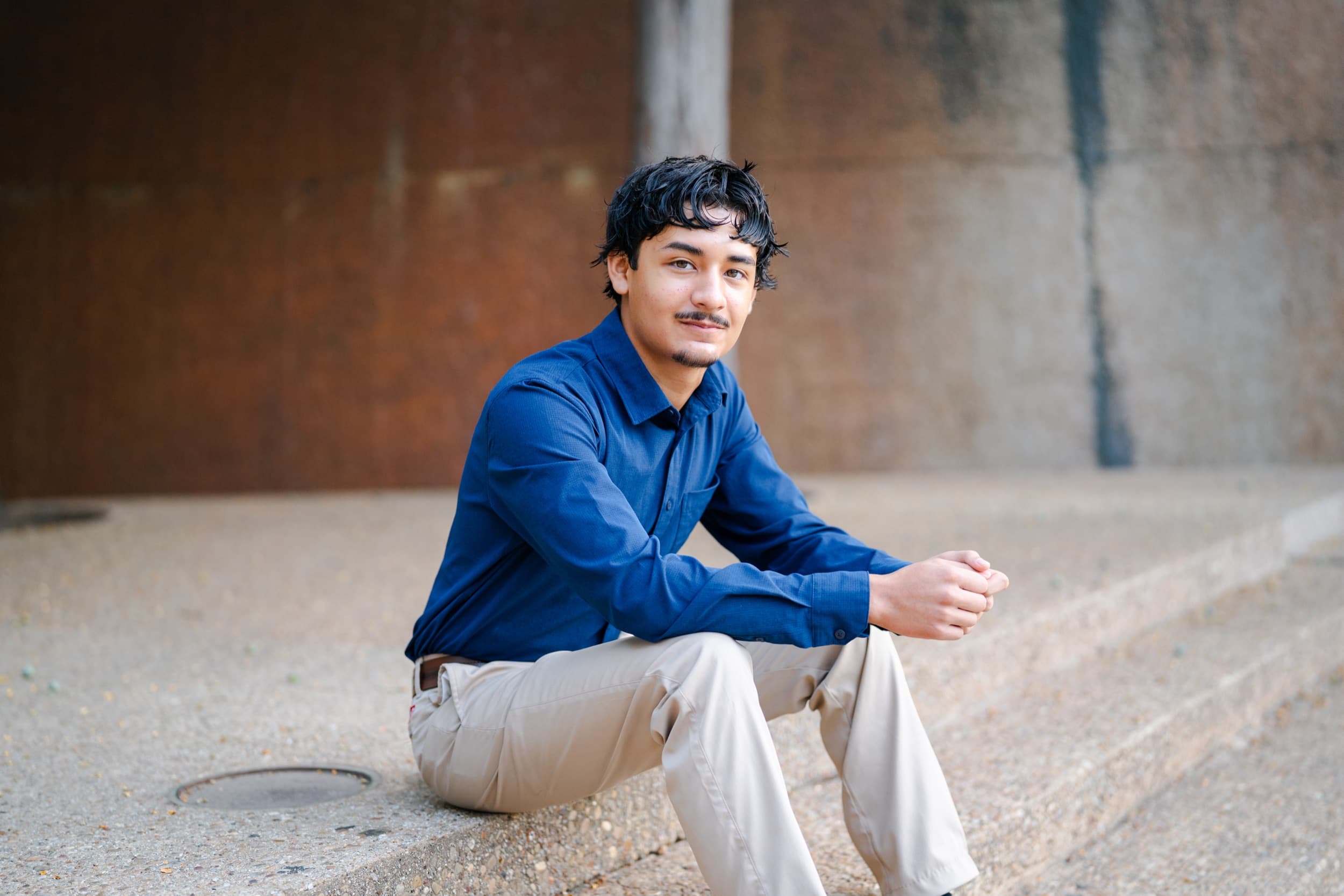 Senior in a navy button-up shirt sitting on concrete steps against a weathered wall.