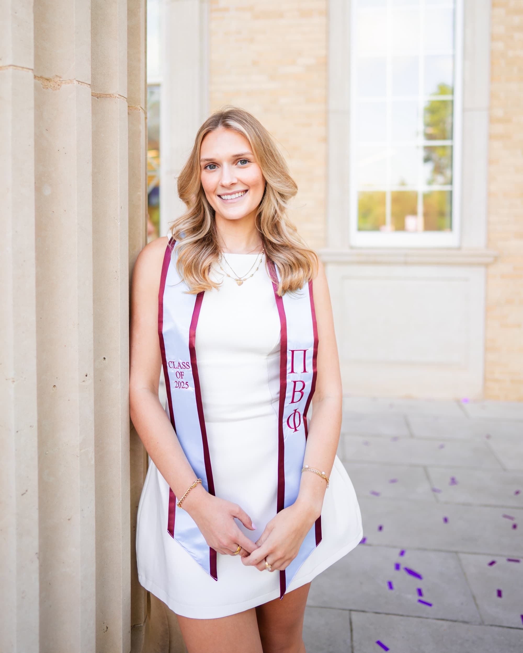Senior in a white dress wearing a graduation stole, leaning against a stone column.