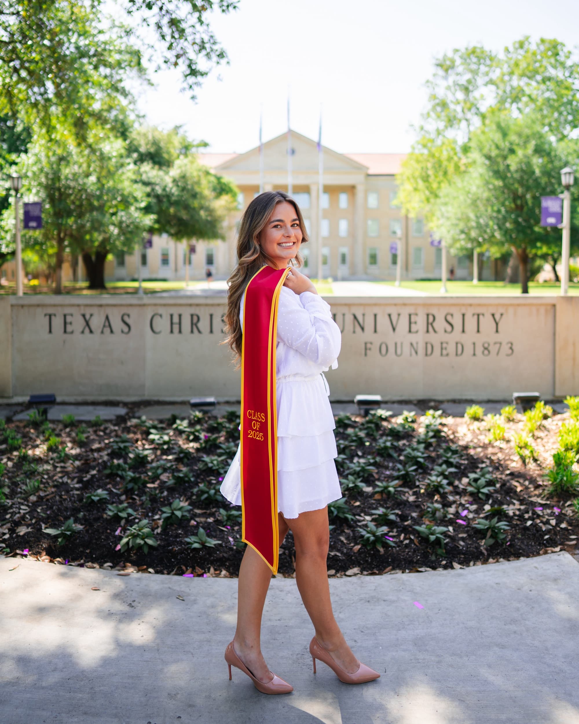 Senior in a white ruffled dress with a red graduation stole draped over her shoulder.