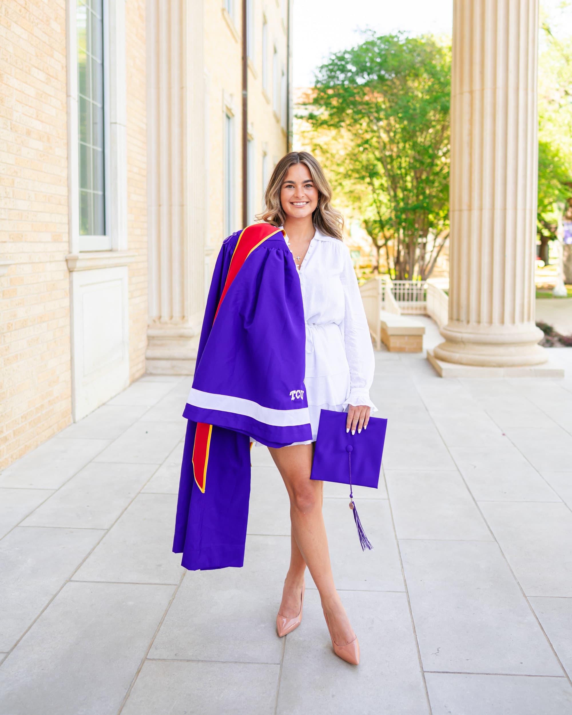Senior in a white dress carrying her purple graduation gown and cap under a colonnade.
