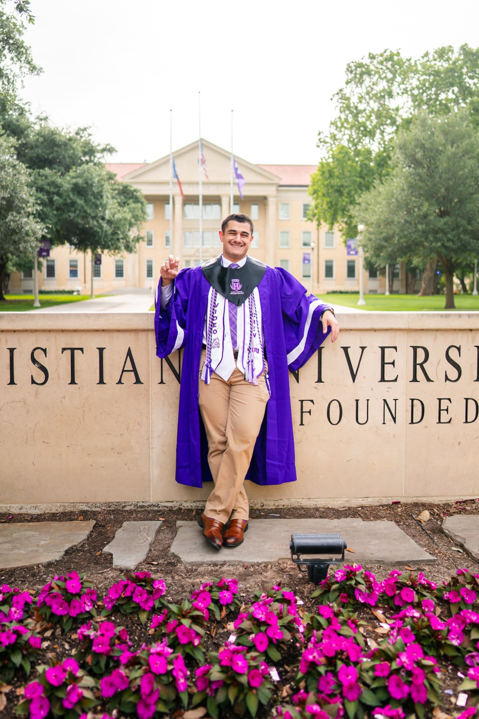 Graduate in a purple honors gown leaning against a university sign beside bright pink flowers.