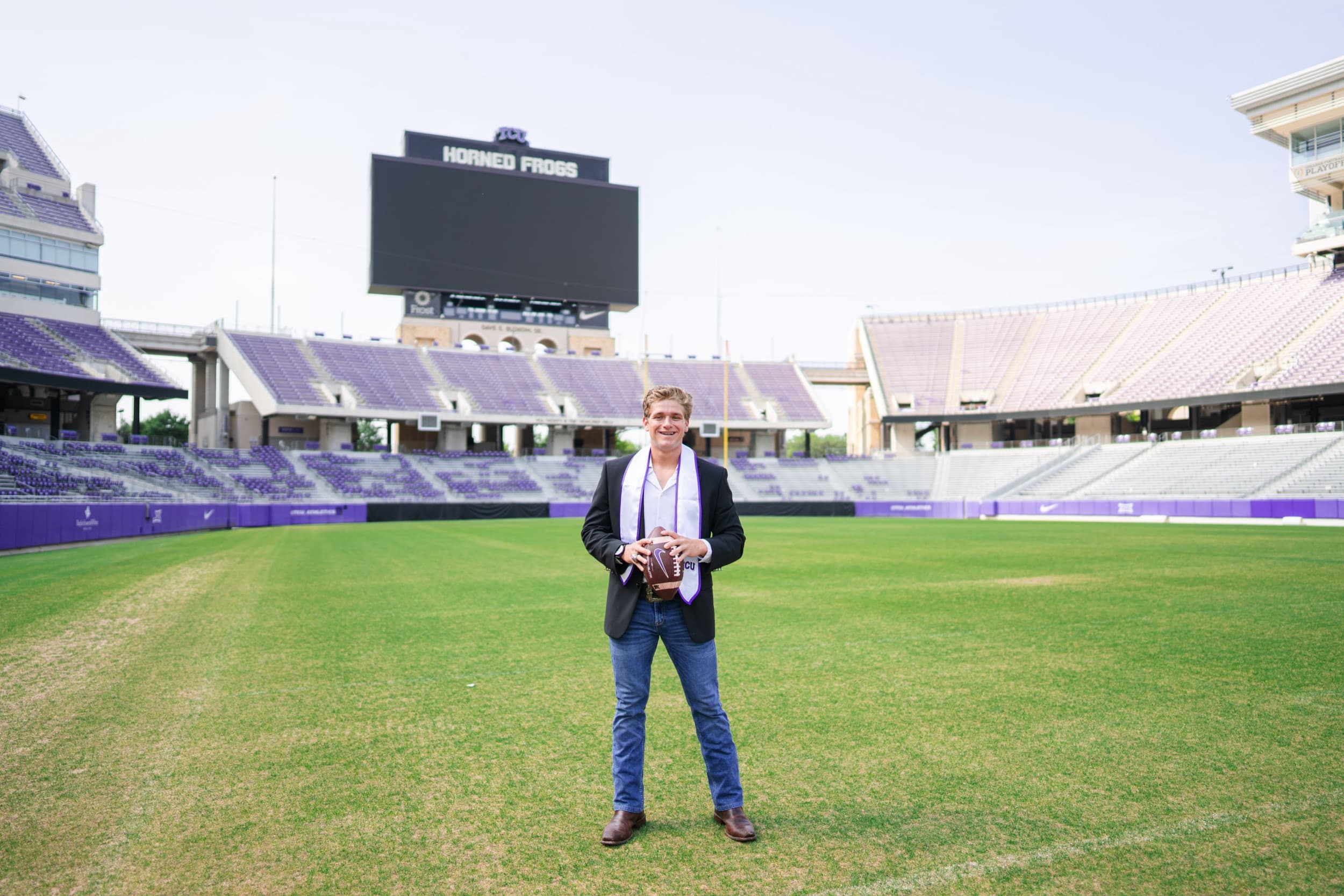 Graduate in a blazer holding a football on the field of an empty stadium.