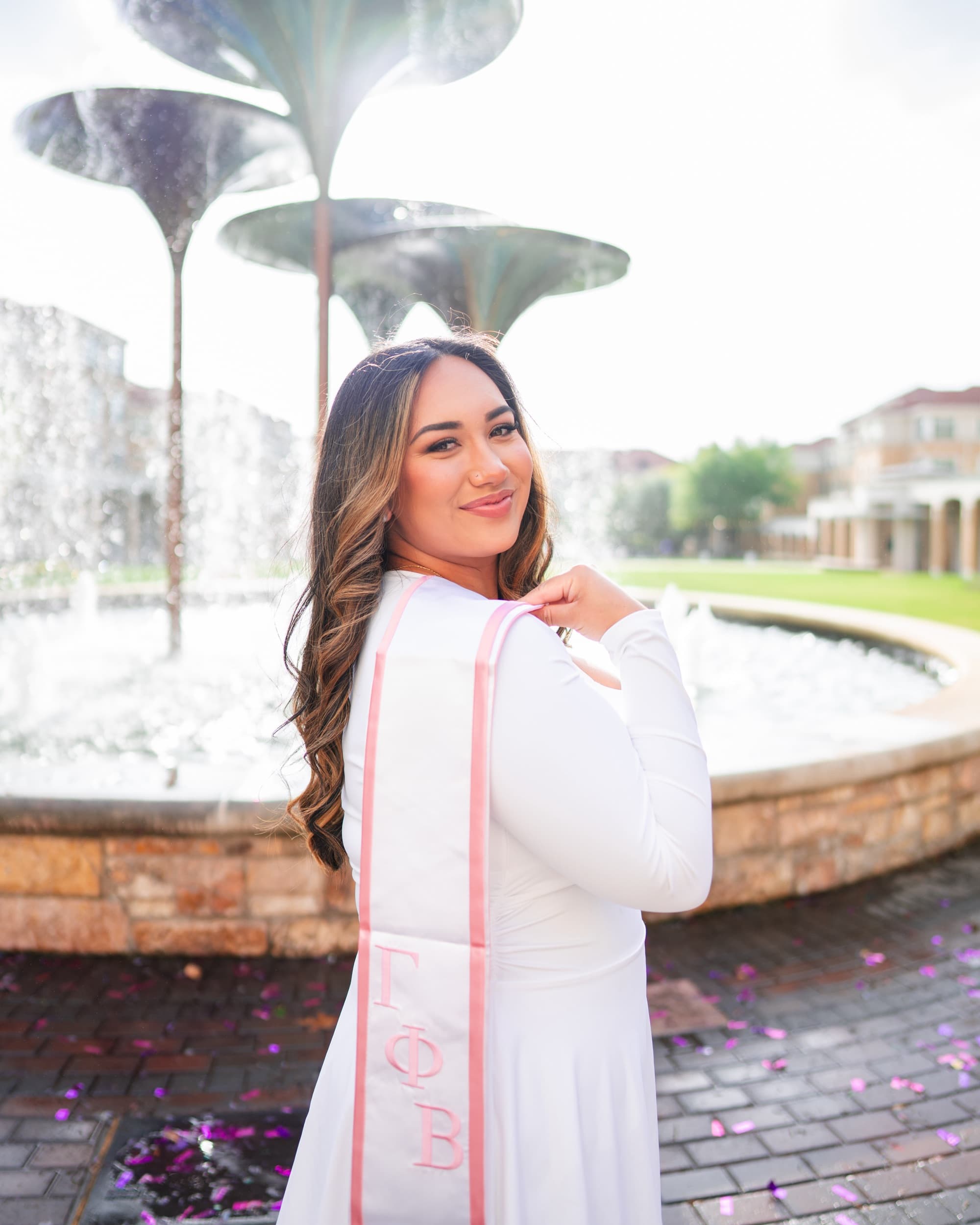 Senior in white glancing over her shoulder, pink graduation stole draped down her back.