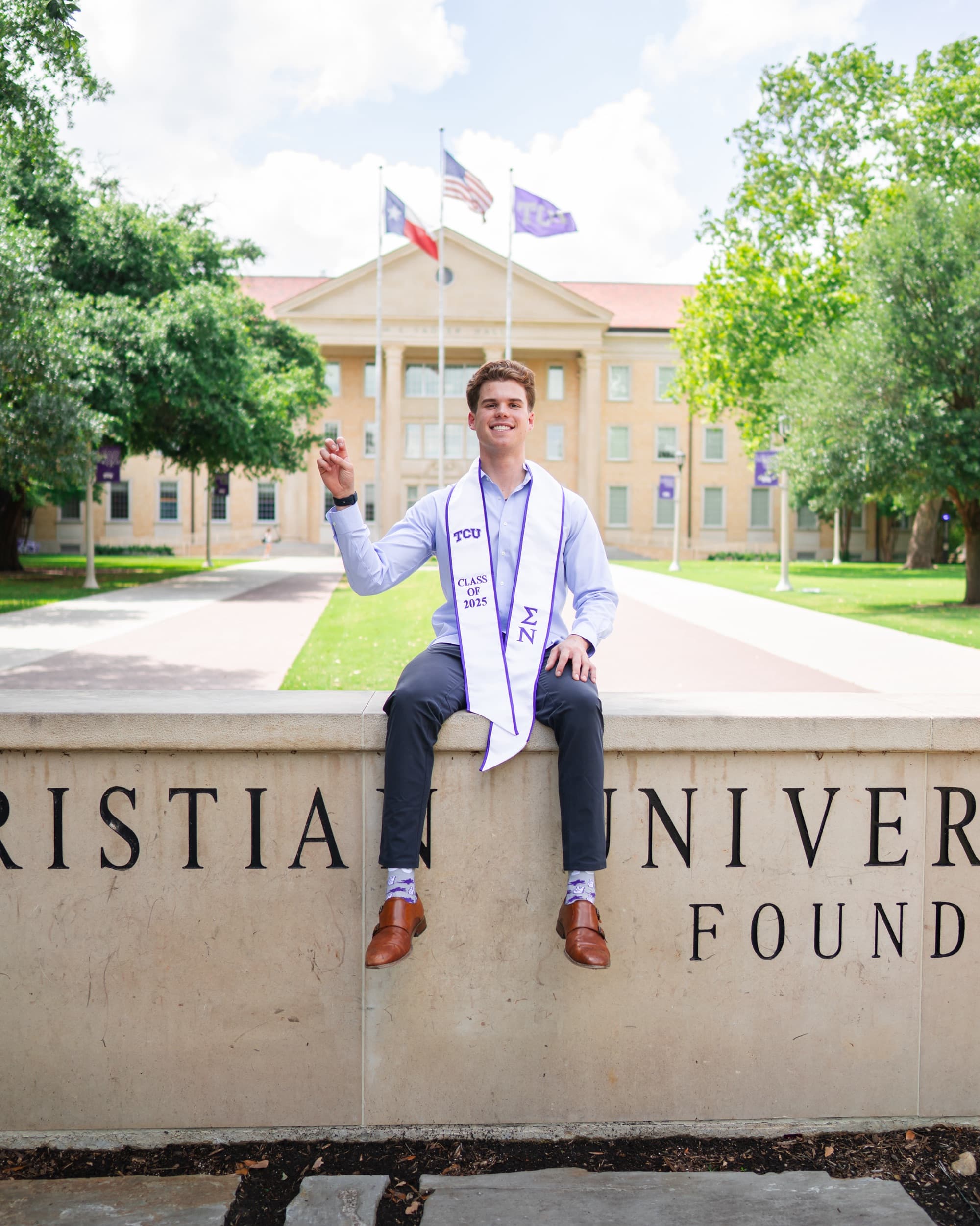Graduate in a graduation stole sitting on a university sign wall, hand raised.