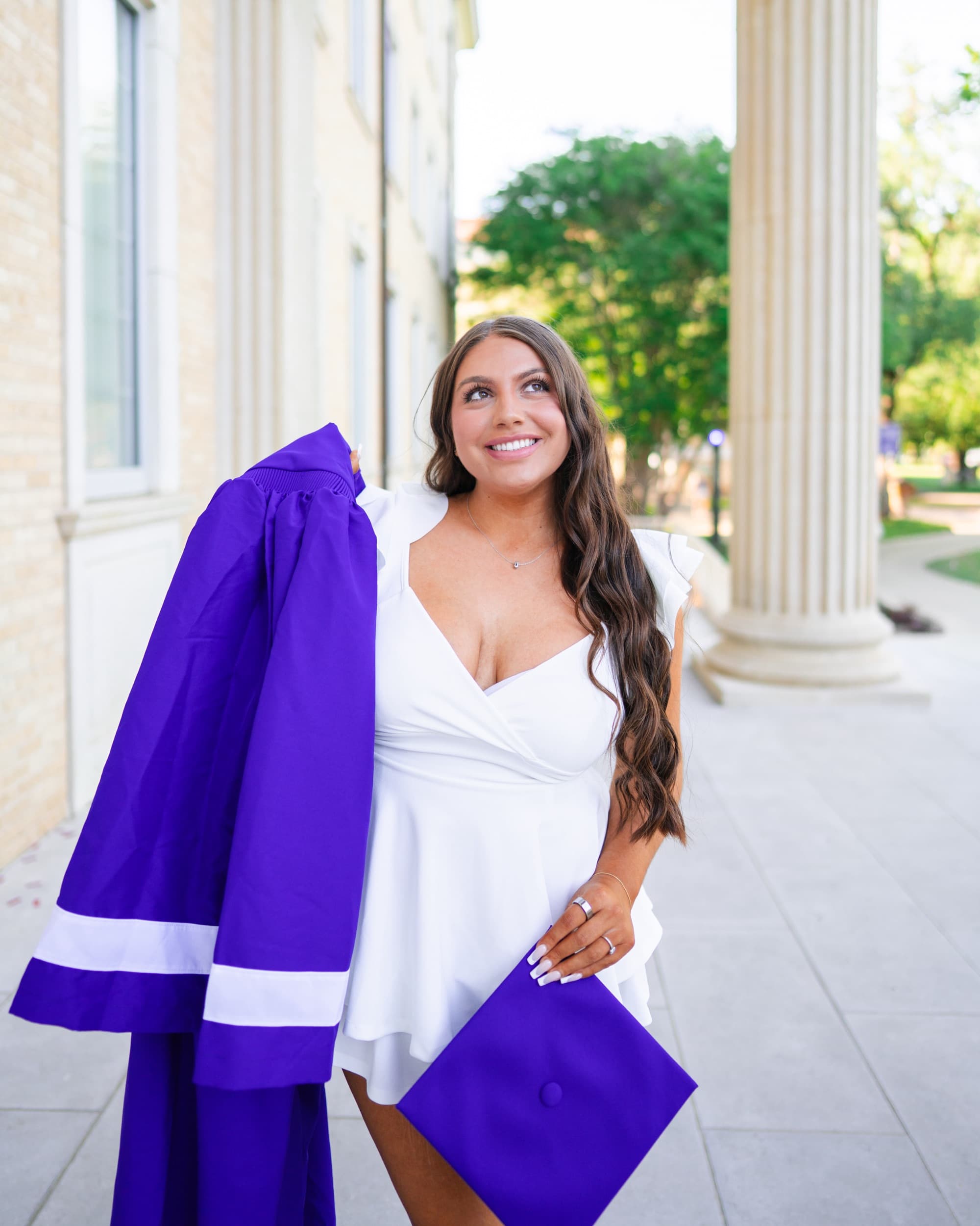 Senior in a white dress glancing upward while holding her purple graduation gown and cap.