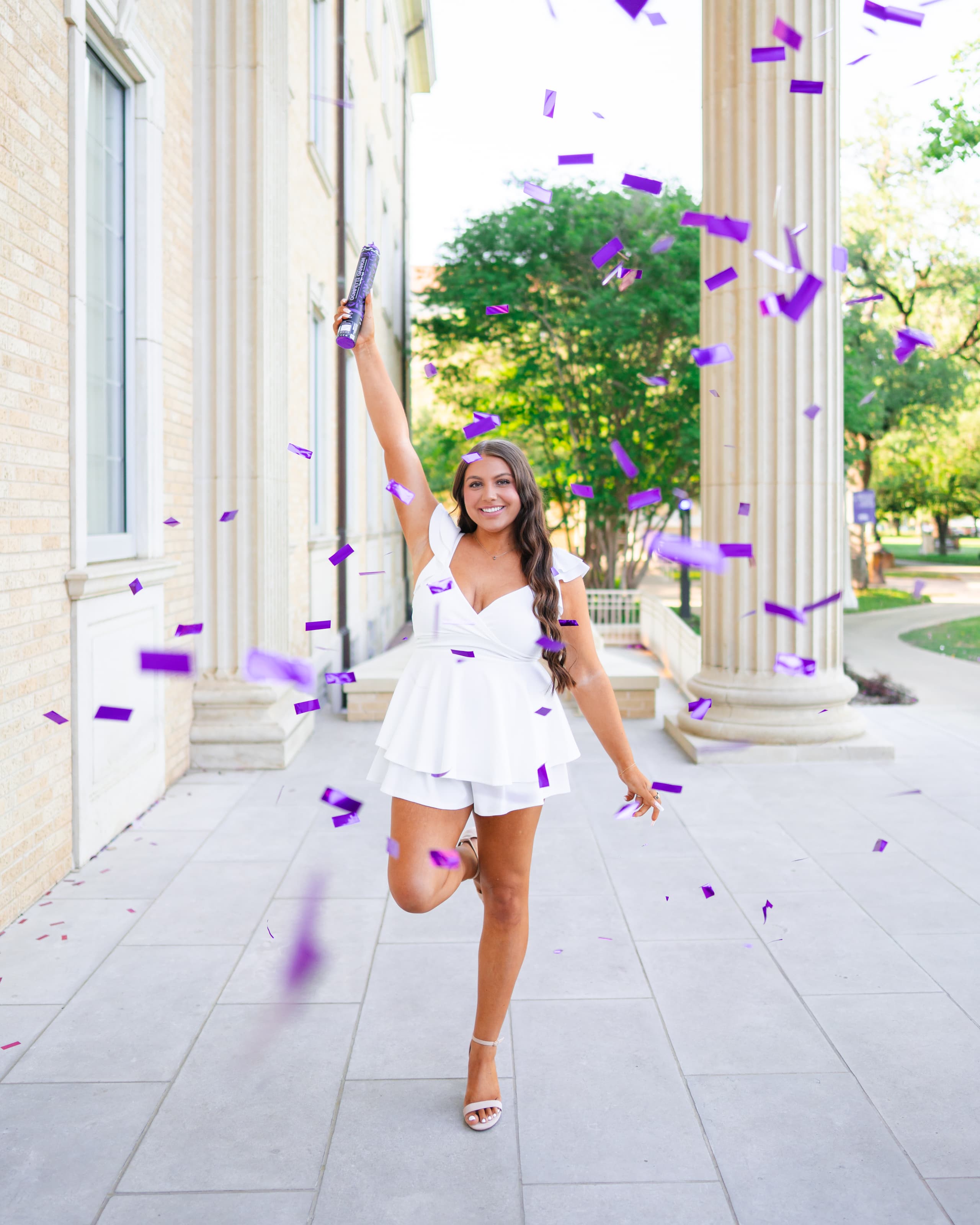 Graduate in a white dress raising a confetti popper, purple confetti falling around her near stone columns.