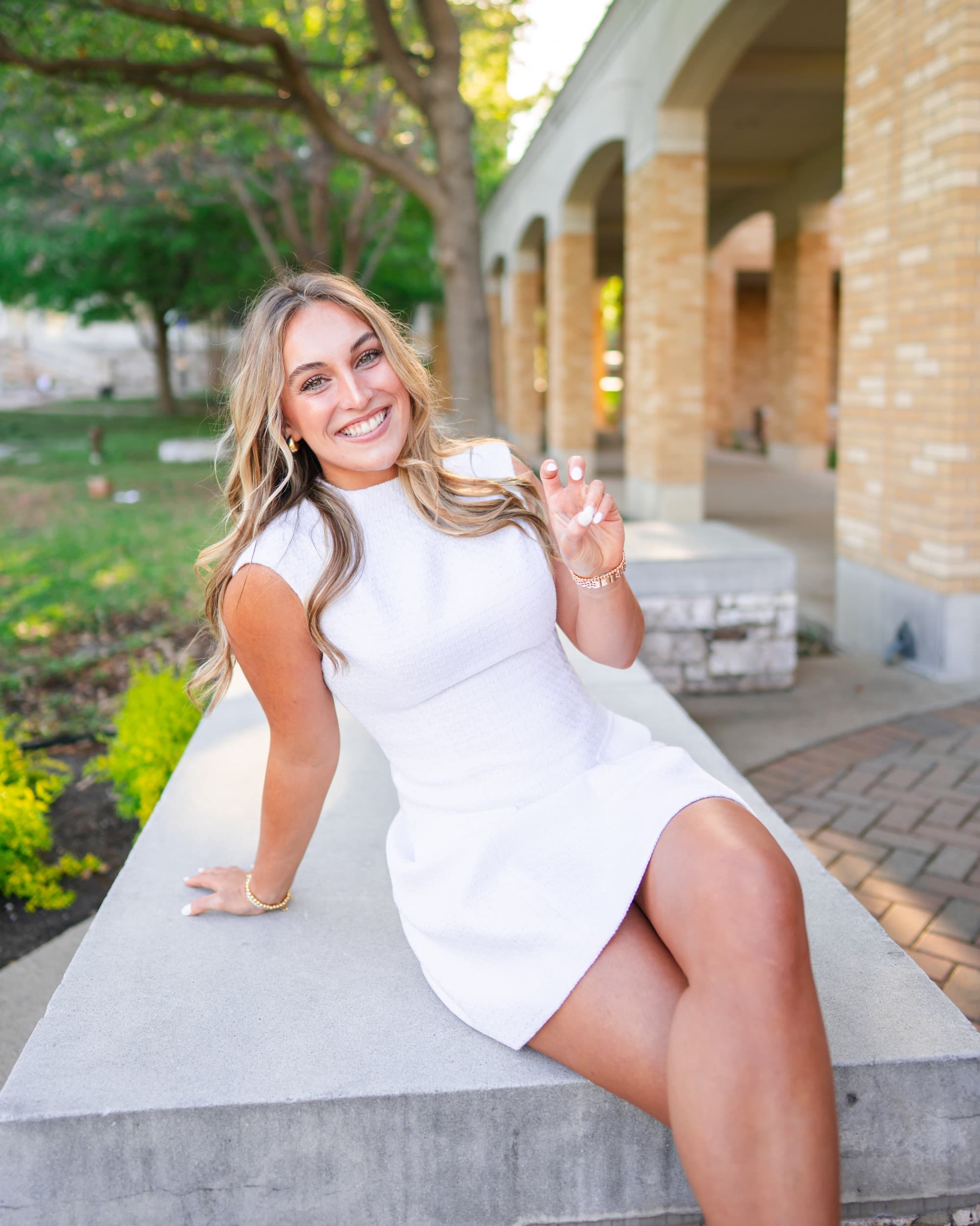 Senior sitting on a stone ledge, smiling brightly beside an arched brick colonnade.