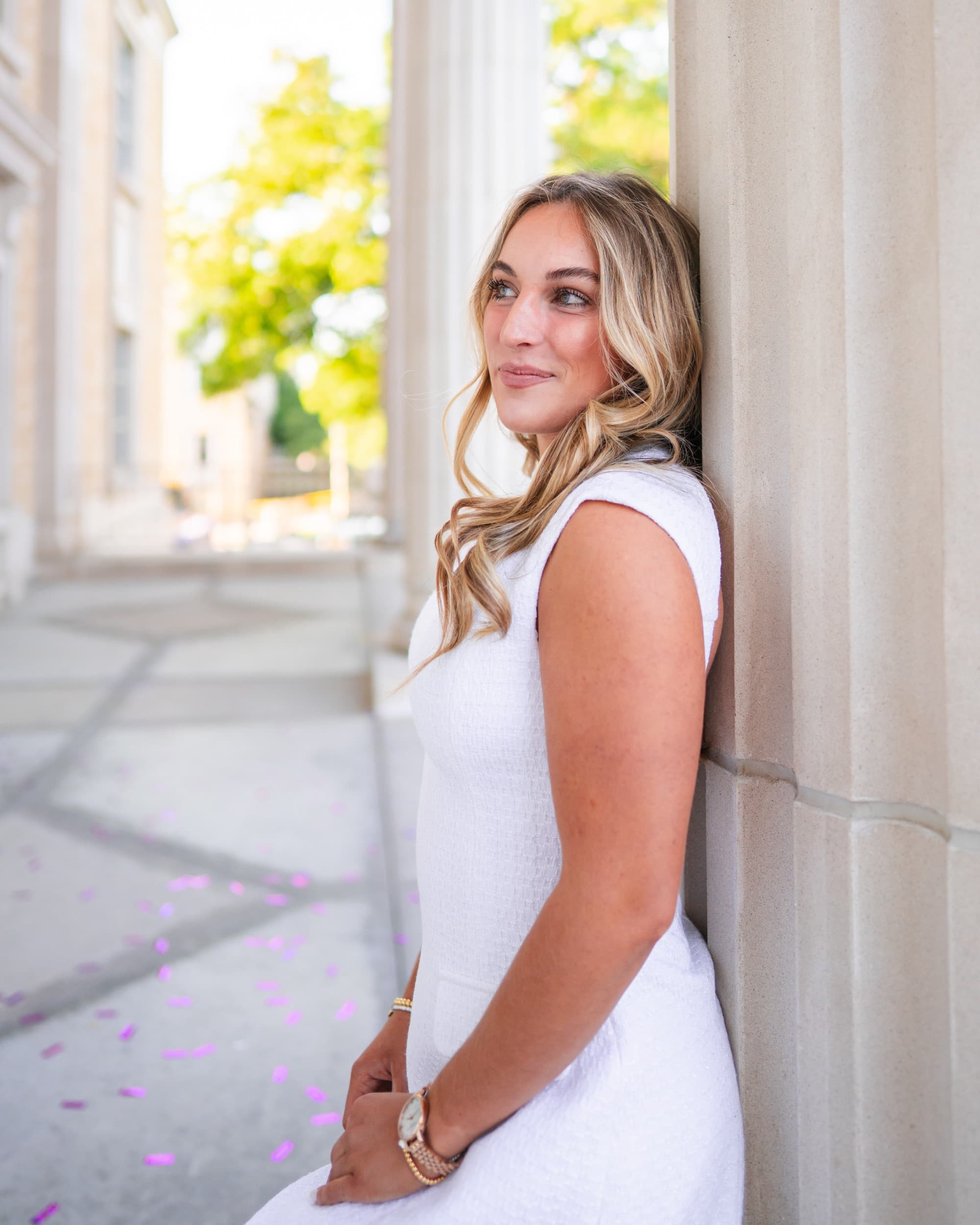 Senior in a white dress leaning against a column, gazing softly into the distance.