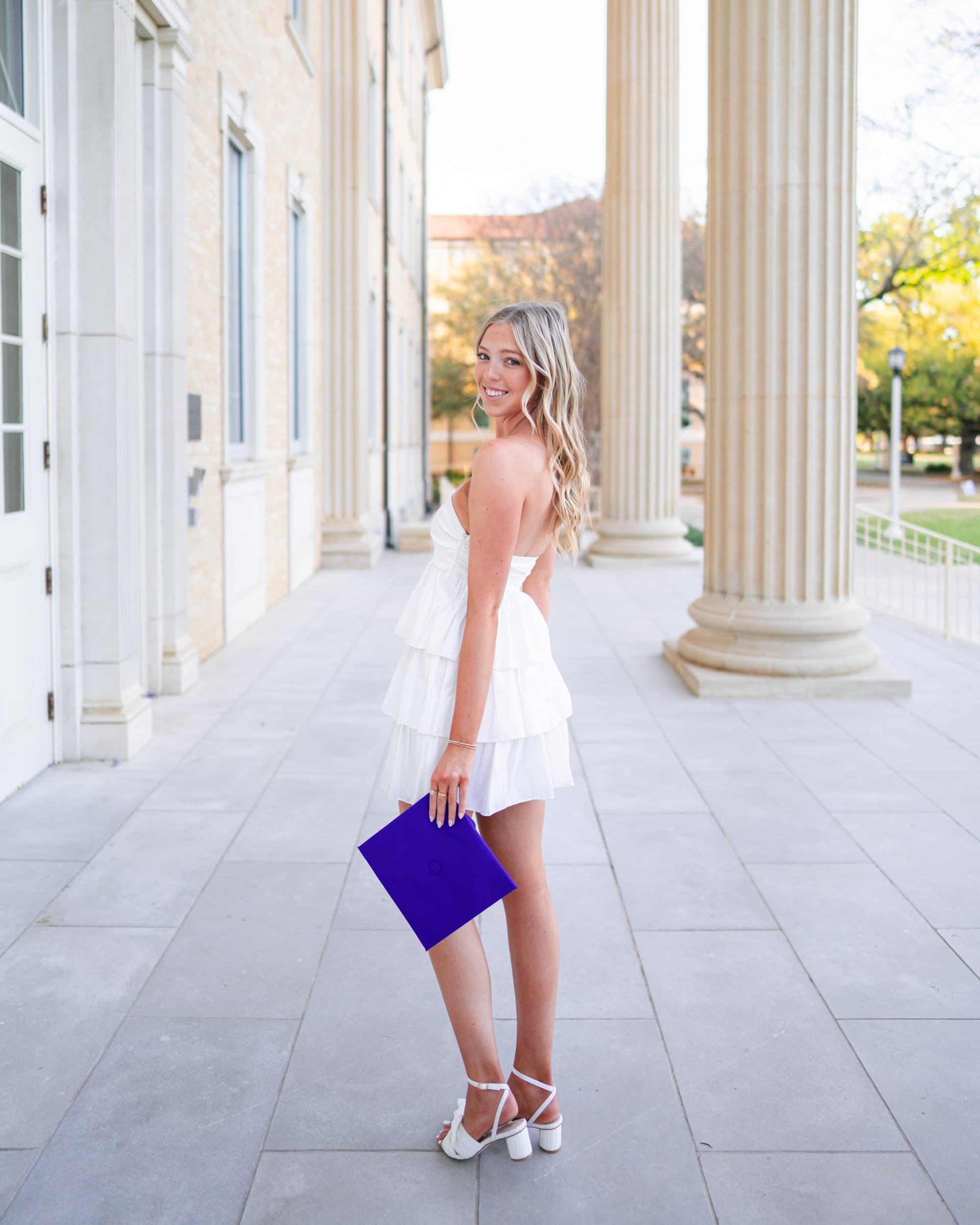 Senior in a tiered white dress glancing back while walking between tall stone columns.