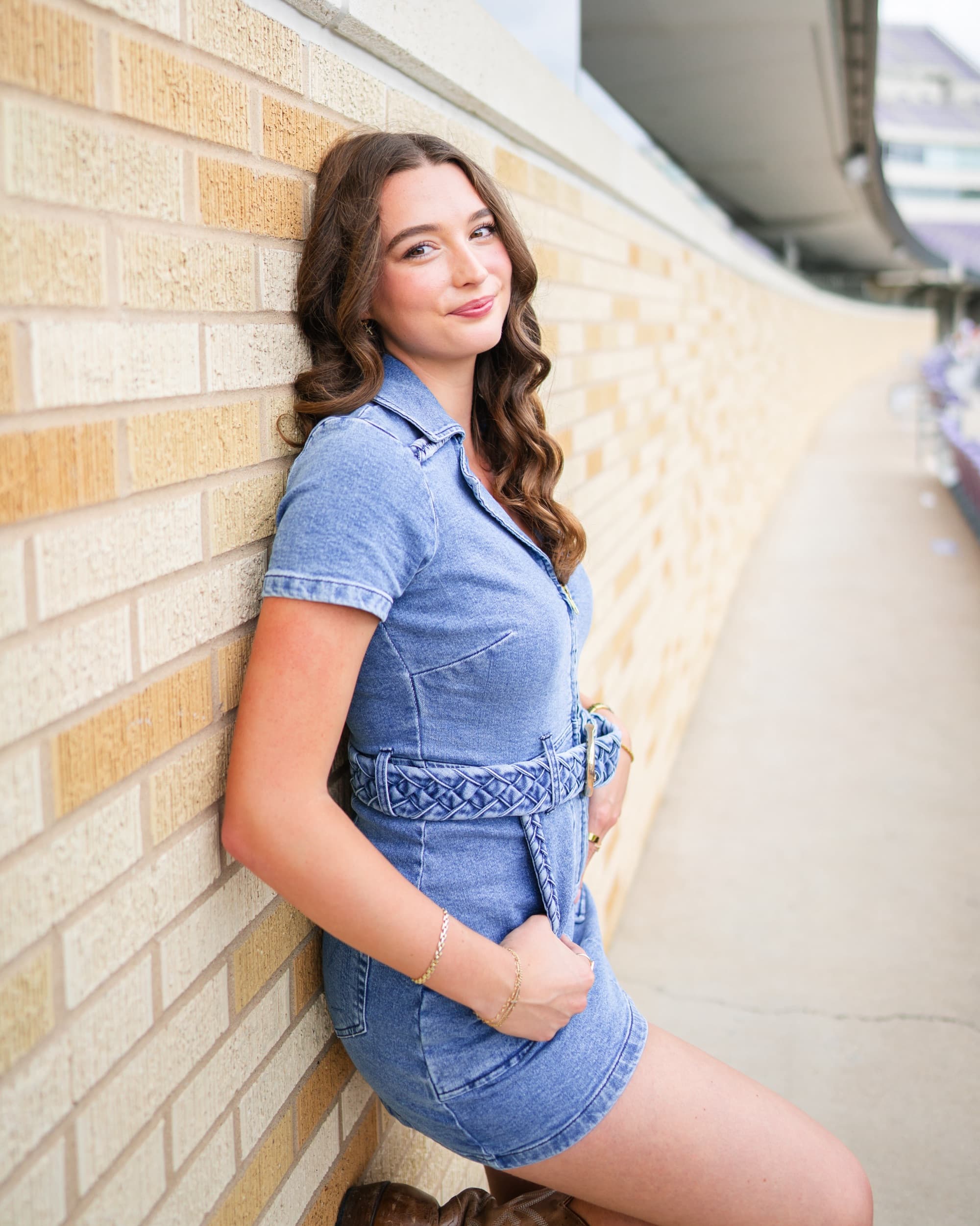 Senior in a denim romper leaning against a brick wall under a stadium overhang.