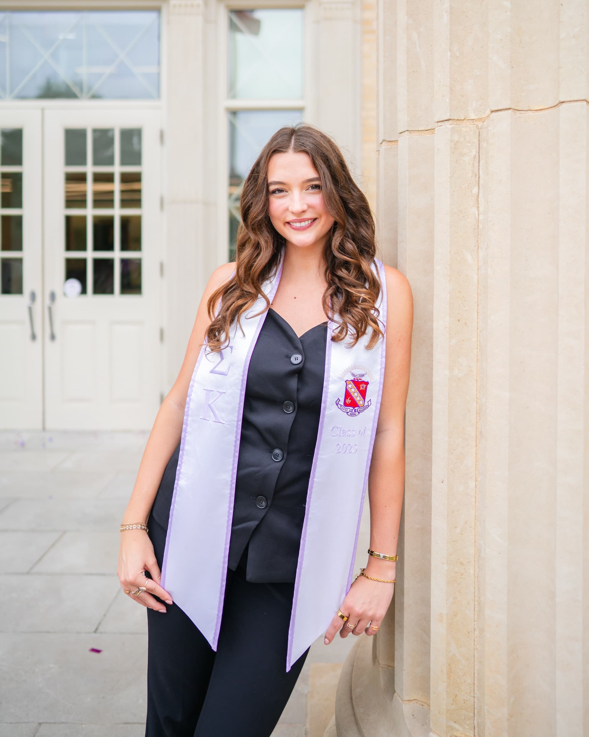 Senior in a black jumpsuit and lavender stole leaning against a carved stone column.
