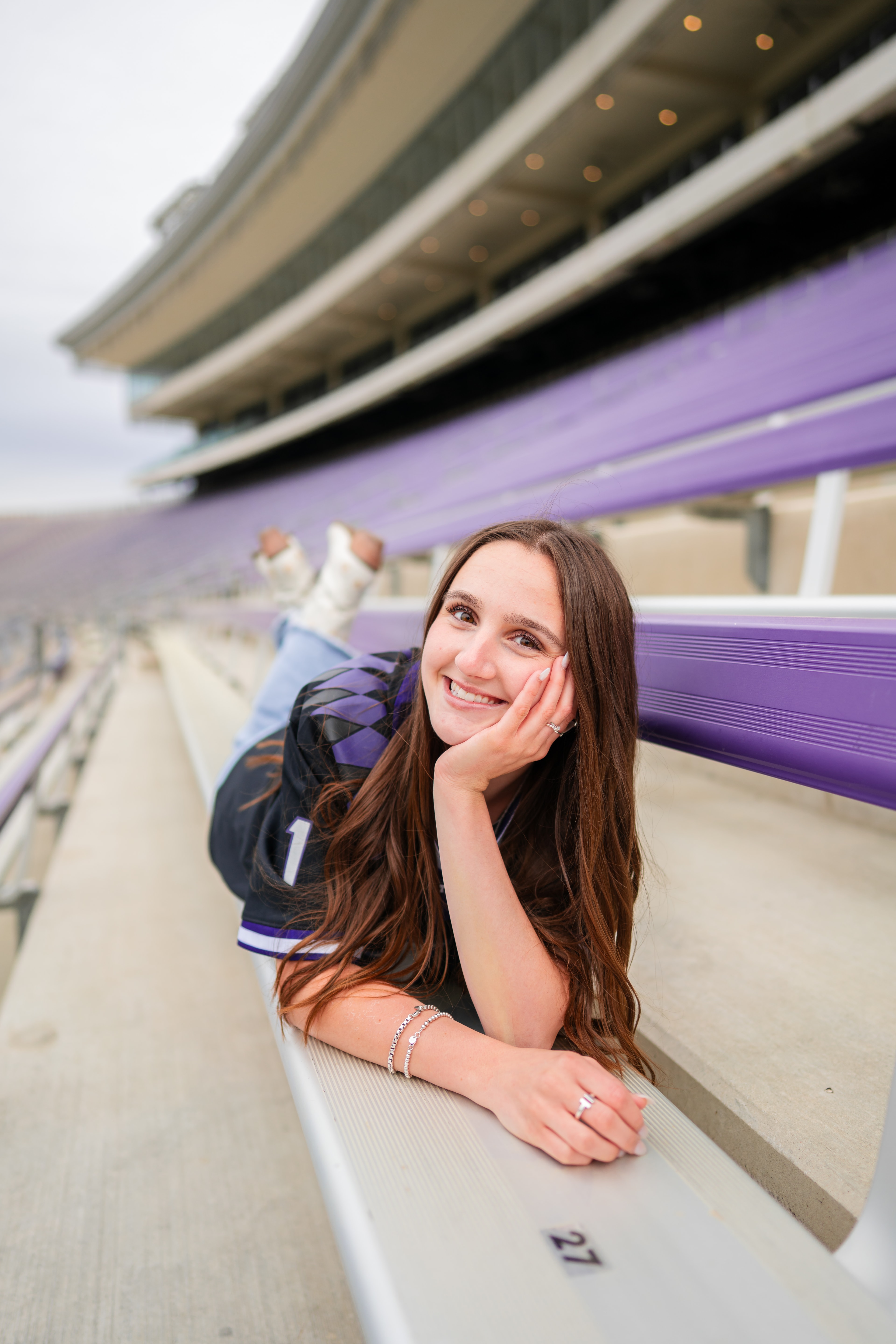 Senior lying playfully on stadium bleachers, chin resting in hand with a bright smile.